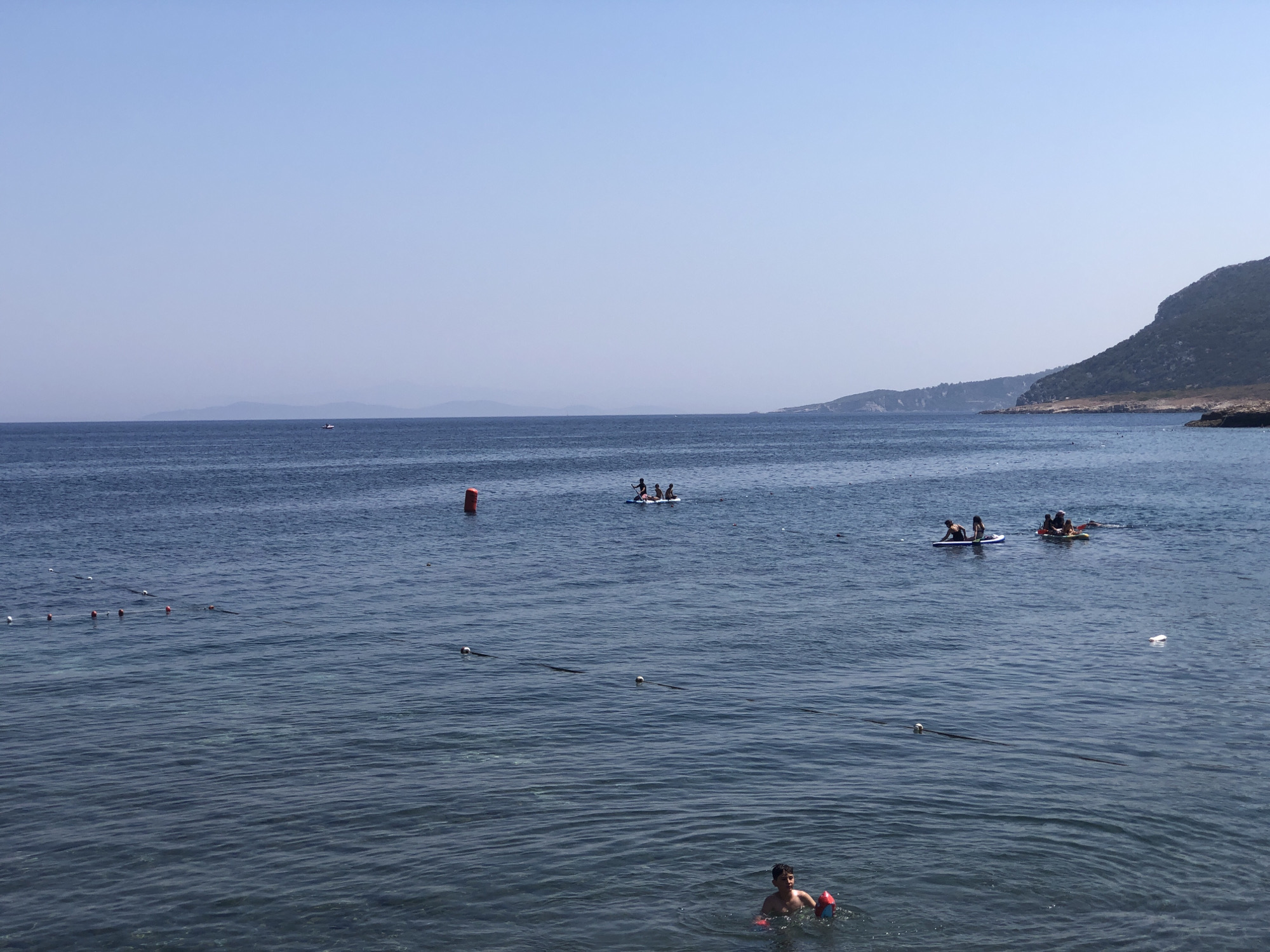The image depicts a tranquil scene at the sea in Karaburun, Turkey. The water is a deep blue, with gentle ripples indicating a calm day. In the foreground, a young boy is swimming, holding a red and blue inflatable toy. Further out in the water, several people are enjoying recreational activities on paddleboards. Two paddleboards are visible, one with three individuals and another with two. A large orange buoy floats in the distance, marking a boundary in the water, and a line of smaller buoys stretches across the left side of the frame, likely designating a swimming area. In the background, the hilly coastline of Karaburun is visible, covered in lush green vegetation, with a clear, pale blue sky overhead. The overall impression is one of a peaceful summer day by the sea.