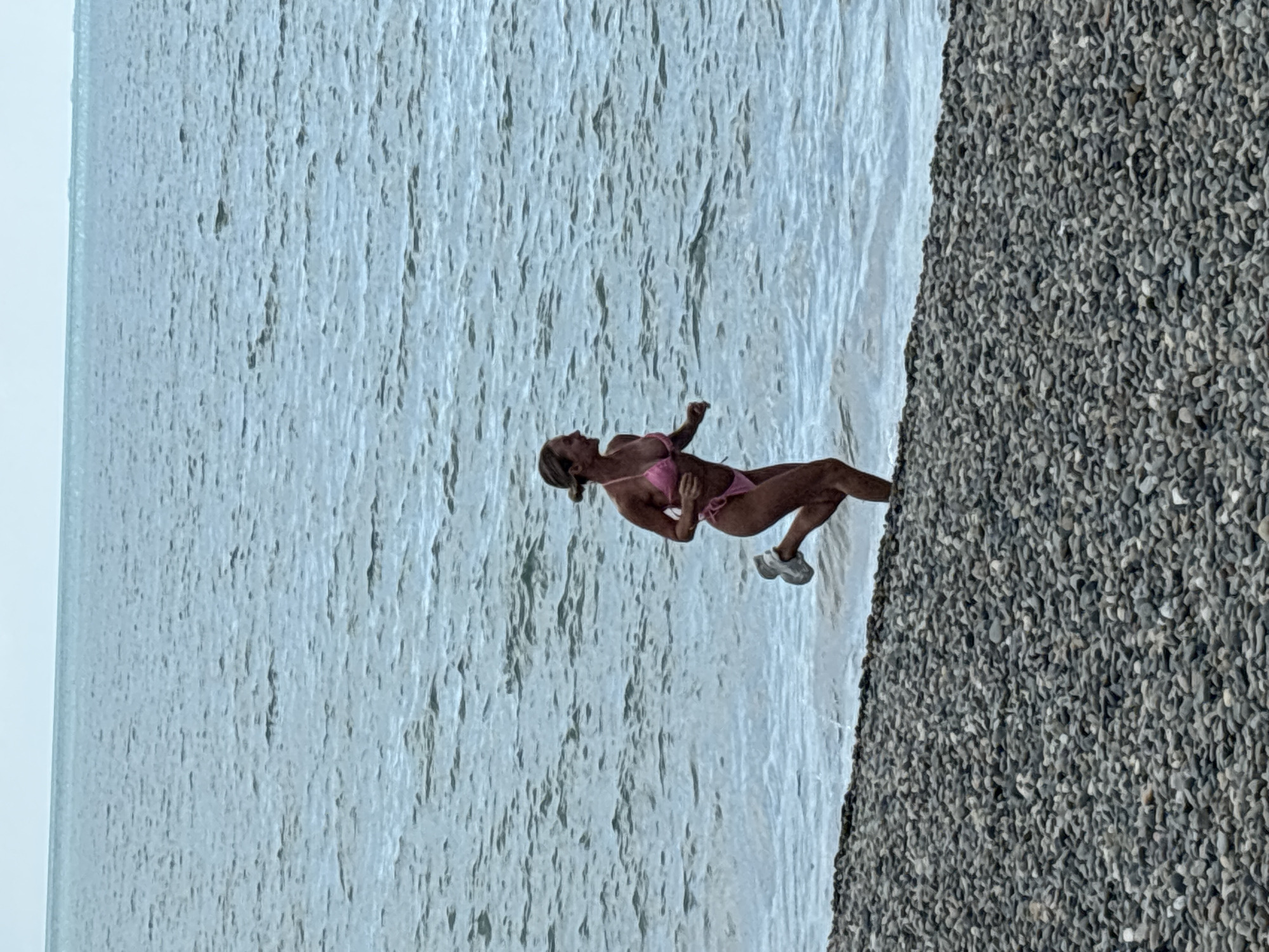A woman in a pink bikini is running on a rocky beach near the water's edge. The sea is calm with light waves. The sky is overcast. The location is a beach in Sochi, Russia.