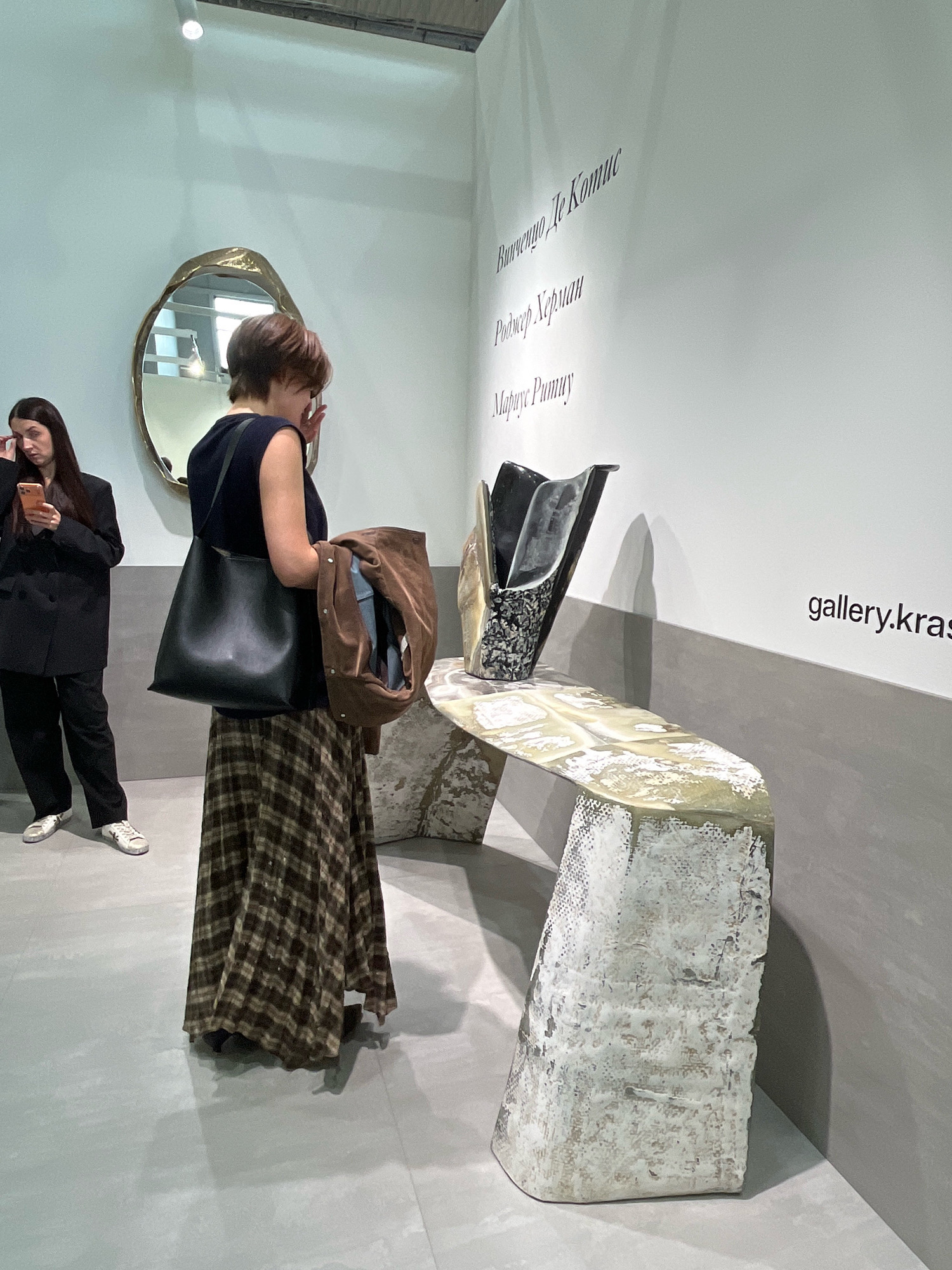 The image shows an indoor scene, likely an art gallery or exhibition, possibly in Khimki, Russia. In the foreground, a woman with short brown hair, wearing a dark sleeveless top and a long plaid skirt, stands with her back mostly to the camera. She is holding a brown jacket over her arm and appears to be touching her face with her right hand. She carries a large black tote bag over her left shoulder. To her left, another woman with long dark hair, wearing a black blazer and black pants, is looking at her phone, with her right hand near her face.
In the background, a large, ornate gold-colored mirror with an irregular shape is mounted on a white wall. The wall also features text in a script font, with some lines of text visible above a contemporary sculpture. The sculpture is displayed on a textured, light-colored console table that has a rough, almost stone-like base. The console table itself has a reflective, marbled surface. To the right of the sculpture, the text "gallery.kras" is visible on the wall. The flooring is a light gray, possibly concrete or polished stone. The lighting appears to be overhead track lighting.