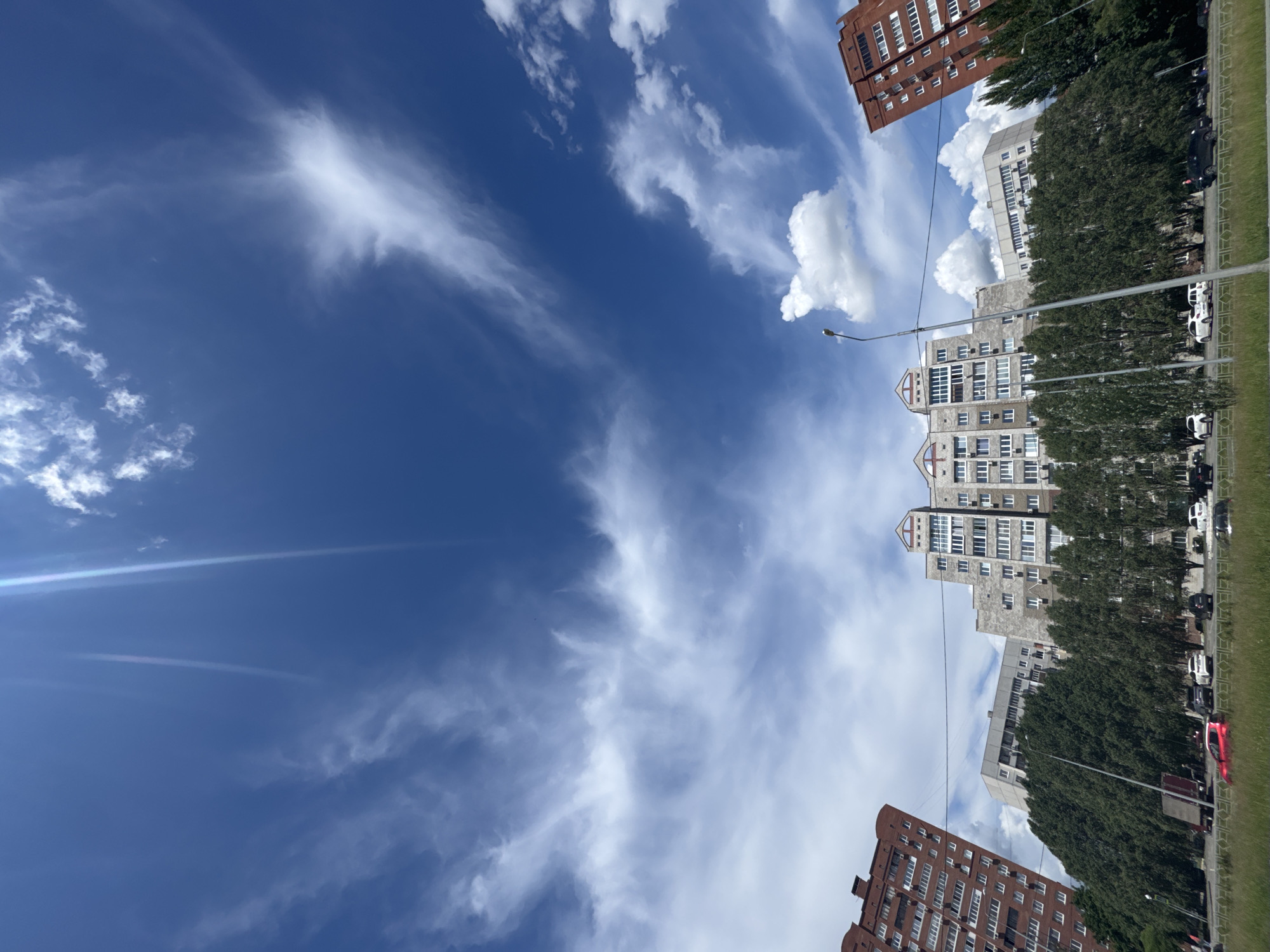 The image presents a view of a city scene, with a blue sky filled with scattered cirrus clouds dominating the upper left portion. The cityscape is visible in the lower right portion of the image. Several multistory buildings of varying heights and colors (brown and light grey) are apparent, interspersed with dense green trees. A row of parked cars can be observed along the edge of the street beside the buildings. A street lamp is also present. The location is Unknown, Unknown.