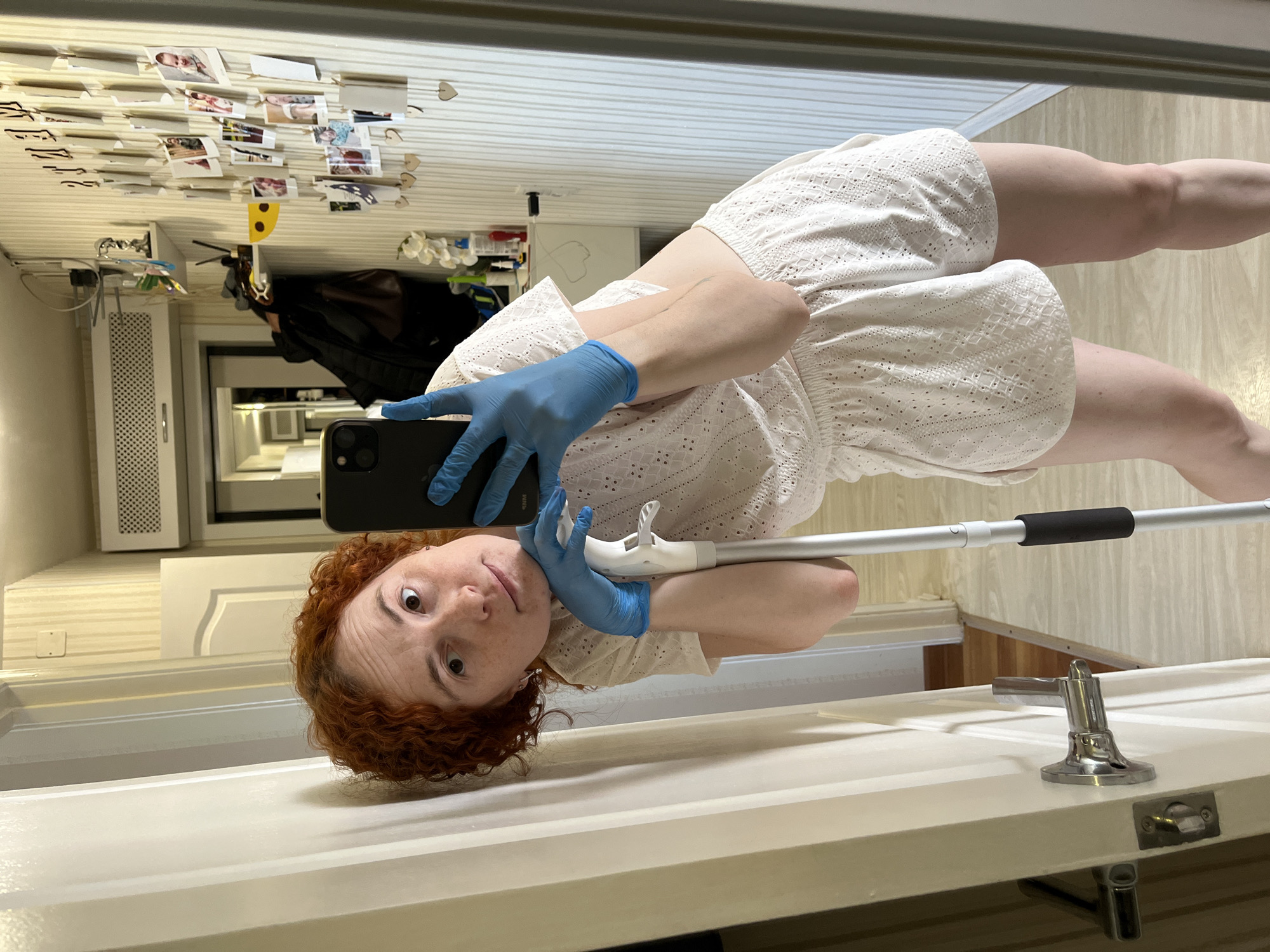 A red-haired woman wearing blue gloves and a white eyelet romper is taking a selfie in a mirror. She holds a black smartphone in her left, gloved hand, and rests her right, gloved hand on a mop handle. The mirror reflects a hallway with a white door, white wall paneling, and various items like a radiator, clothing rack with a black garment, and a collection of photographs and paper hearts attached to the wall. A chrome door handle is visible on the lower right side of the frame.
There is insufficient information to determine the location of this scene within the city of Chaikovsky, Russia.