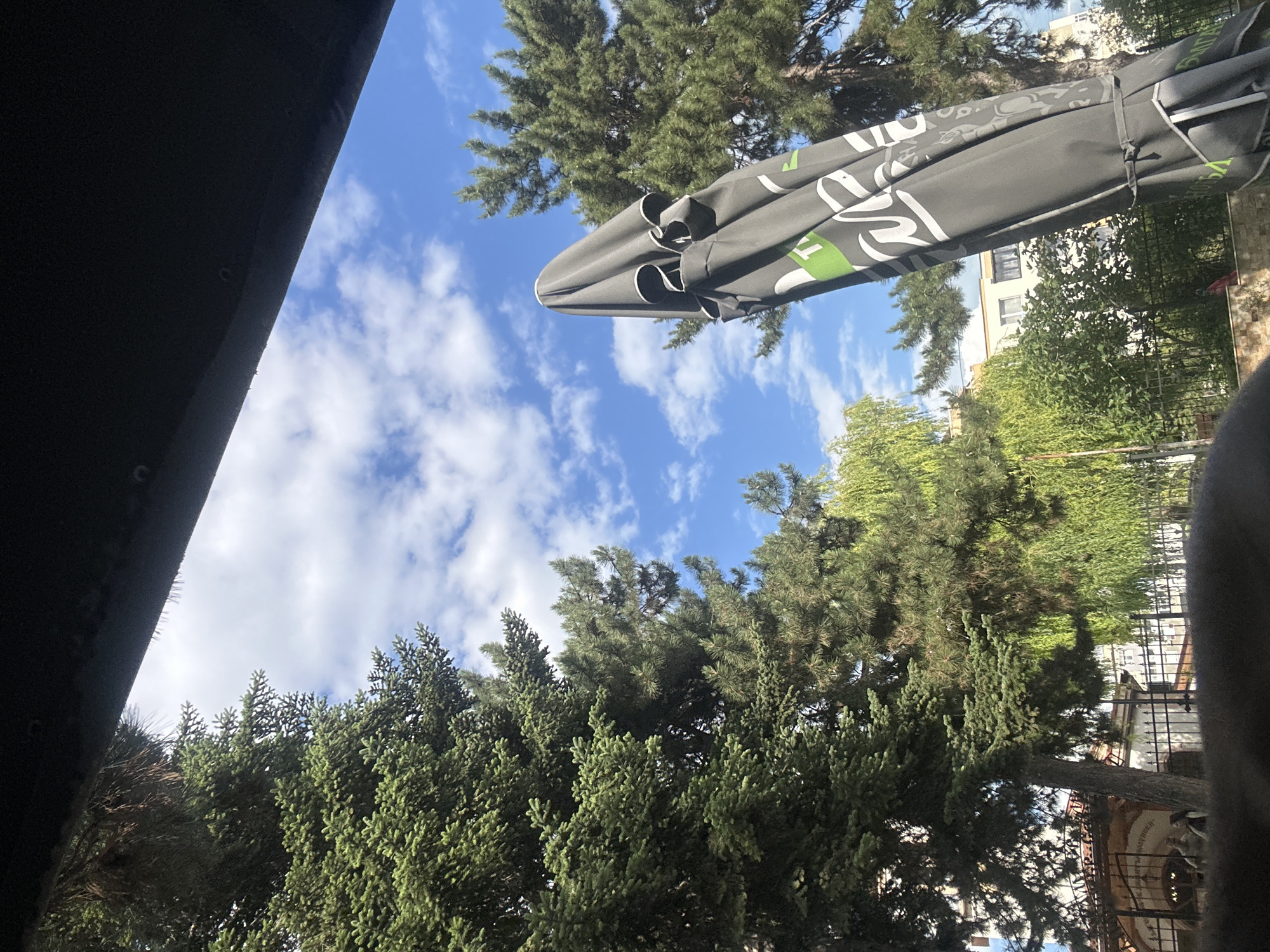 The image depicts an outdoor scene with a focus on a closed gray umbrella in the foreground, partially obscuring a blue sky with scattered white clouds. To the left, a large evergreen tree with dense needles dominates the frame, with another evergreen tree visible to the right, its branches thinner and more spread out. In the background, a building with light-colored walls and windows is visible, situated behind a fence and lush green foliage, including what appears to be a weeping willow tree. The lower portion of the image is obscured by a dark, blurry foreground element, possibly the edge of a structure or furniture. The scene suggests a casual outdoor setting, potentially a cafe or restaurant, in the city of Varna, Bulgaria.