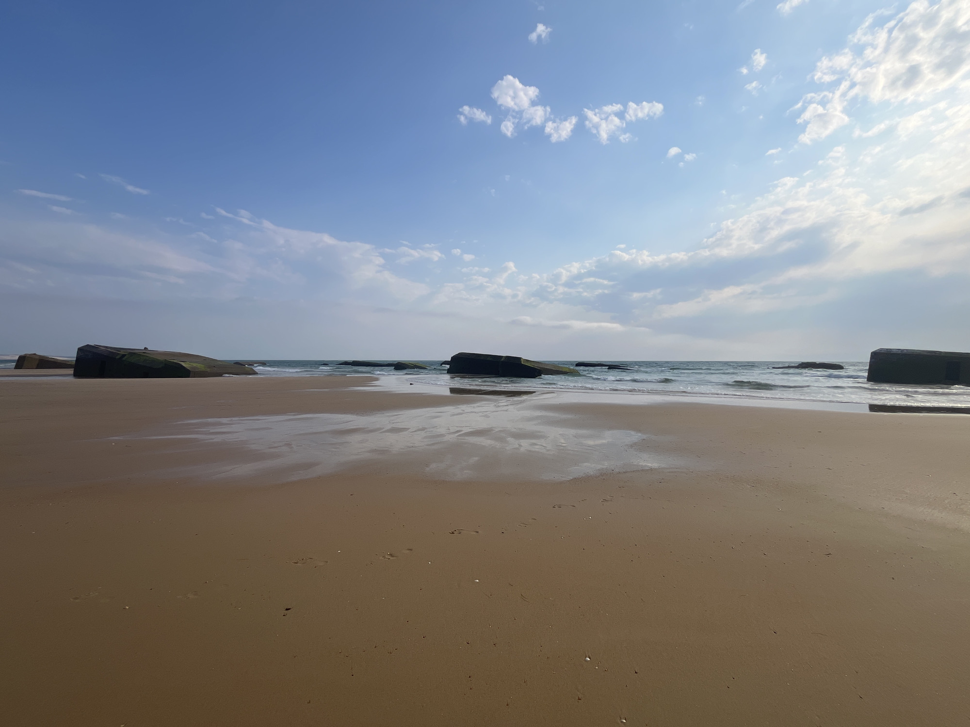 The image shows a sandy beach at low tide in Lège-Cap-Ferret, France, with several concrete bunker remnants protruding from the sand and shallow water. The sky is blue with scattered white clouds. The waves are gentle, lapping against the shore and around the bunkers. Footprints are visible in the wet sand closer to the viewer. The scene is peaceful and serene, with no people present. The bunkers, likely remnants of World War II defenses, are partially submerged in the water and sand, creating a striking contrast between the man-made structures and the natural environment.