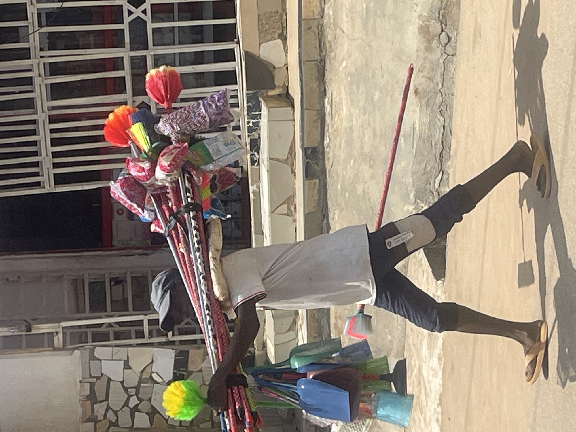 A man wearing a dark baseball cap, a white t-shirt, and dark pants walks in a city street, carrying a bundle of brooms and dusters on his shoulder. The brooms have colorful handles and brush heads, some are red, orange, yellow, and green. Several plastic dustpans, one blue, one green, and one light blue, hang from the man's arm. He is walking from left to right across the frame, casting a shadow on the sandy ground. The background shows a building with a stone facade and a metal grate structure. The scene appears to be in Abuja, Nigeria, given the context provided.
