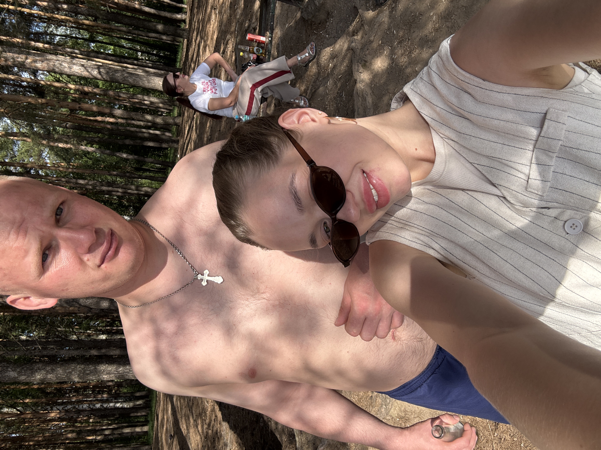 A selfie taken in a wooded area near Chaikovsky, Russia, features a shirtless man with a silver cross necklace and blue shorts, posing with a woman wearing sunglasses and a striped shirt. In the background, another woman is lying on the ground with bottles and cans nearby.