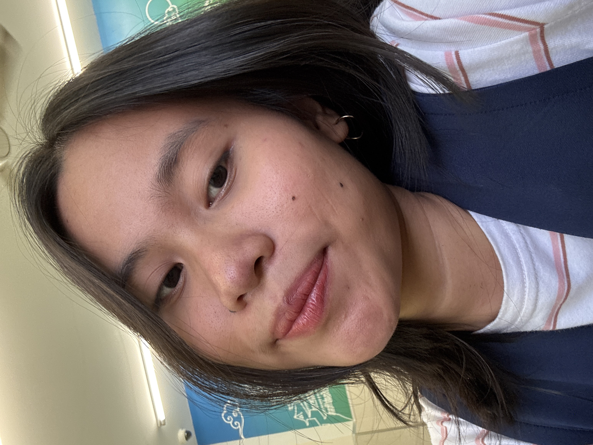 A medium shot, head-and-shoulders selfie of a young East Asian woman is shown in a brightly lit indoor space. She is looking slightly off-camera to her right with a subtle smile. Her dark hair is shoulder-length and slightly windswept. She has dark eyes, visible pores on her skin, a mole above her lip, and two moles on her cheek. She is wearing a white t-shirt with pink and red stripes, and a dark blue vest. A subtle hoop earring is visible in her right ear. In the background, a white ceiling with fluorescent lighting and a portion of a wall with blue and green graphics featuring stylized clouds and a building can be seen. The overall impression is casual, possibly taken in a school or public building.