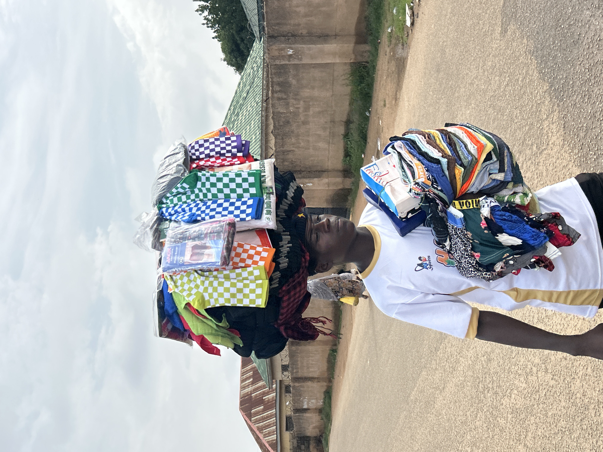 In Jos, Nigeria, a young male street vendor walks down a paved road, carrying a large assortment of folded clothing items on his head, with another stack of clothes held in his arms. He wears a white t-shirt with a yellow collar and trim, and black shorts. The clothing on his head is diverse, featuring checkered patterns in blue, green, red, and purple, as well as solid colors and some items in plastic packaging. The clothing in his arms includes various colors and patterns, some of which are labeled with text. The background shows a concrete wall, a building with a corrugated iron roof, and trees. The sky is overcast with clouds. The road appears to be a residential or commercial street.