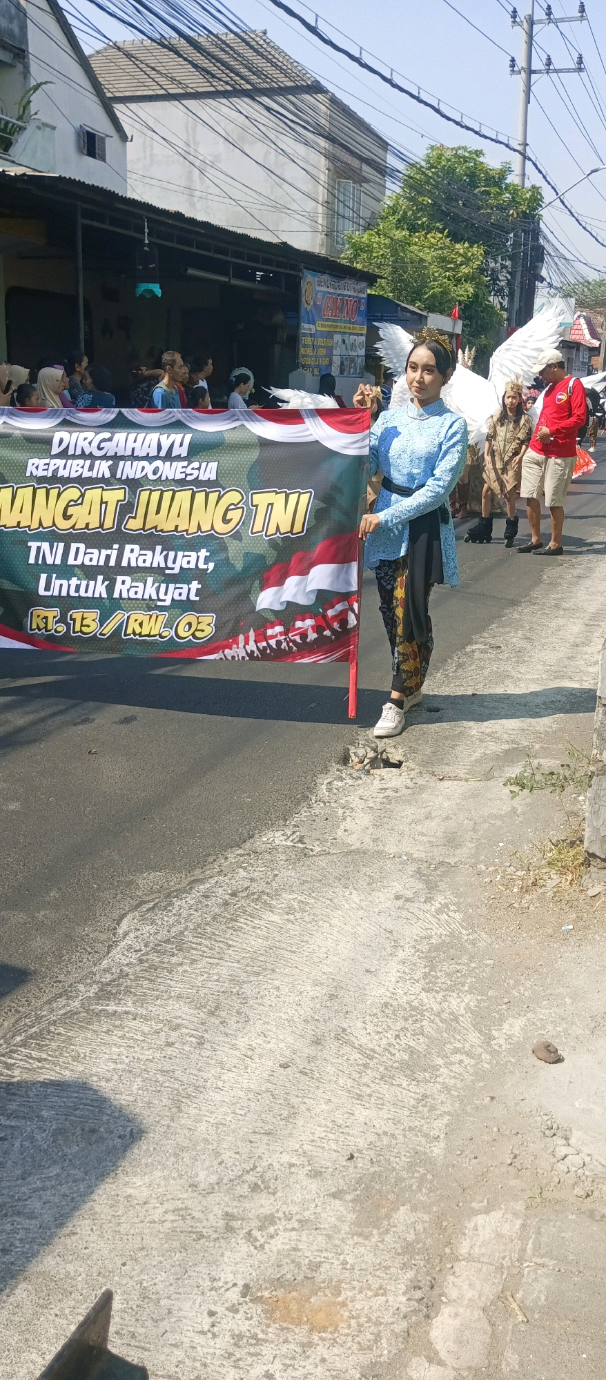 An outdoor, mid-shot, eye-level photograph captures a procession on a street in the city of Malang, Indonesia, under a bright, clear sky. A young woman, dressed in a light blue, traditional Indonesian top and patterned pants, stands on the right side of the frame, holding the left side of a large, olive-green banner. She wears a golden crown and large white feathered wings, suggesting a costume or performance. The banner displays text in white and yellow lettering, including "DIRGAHAYU REPUBLIK INDONESIA" and "MANGAT JUANG TNI," along with an image of the Indonesian flag. Behind her, other participants in the procession are visible, including a person in a brown costume with wings, and a man in a red shirt and khaki shorts. In the background, buildings with tiled roofs and electrical wires line the street, typical of a residential or commercial area in Indonesia. The street itself is paved with asphalt, showing some cracks and a drain on the right side. The overall atmosphere is festive, likely part of a cultural event or parade.
