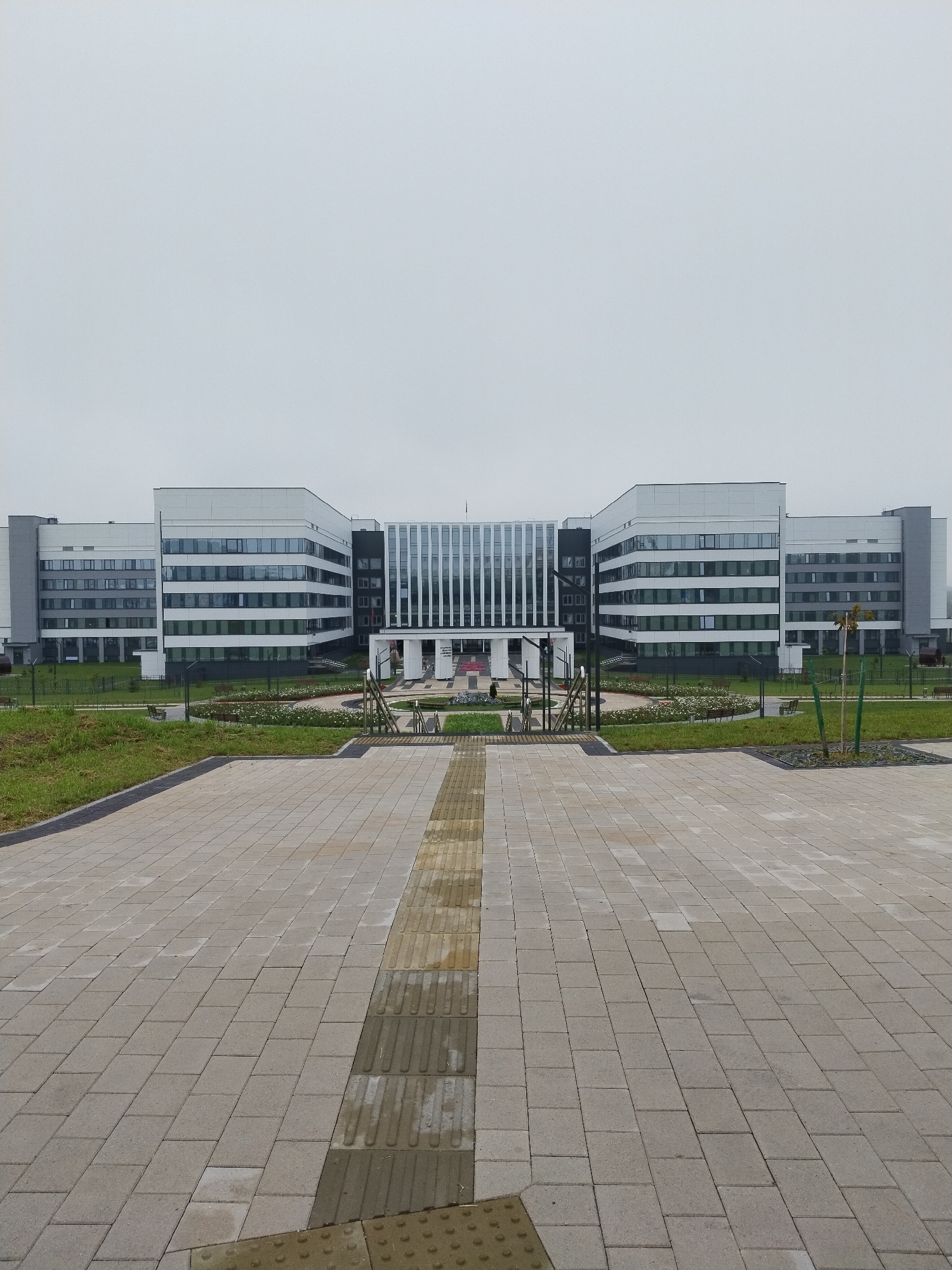 The image depicts a wide, modern, multi-story building with a symmetrical facade composed of white and grey panels and numerous windows. The building appears to be an institutional or commercial structure. In front of the building is a paved plaza with a textured strip running down the center, likely a tactile paving for the visually impaired. Beyond the plaza, there is a manicured green space with flower beds, benches, and a few young trees. The sky is overcast and uniformly grey. The scene appears to be a public area, possibly in front of a hospital, university, or government facility, located in the city of Hrodna, Belarus. There are no people visible in the image.