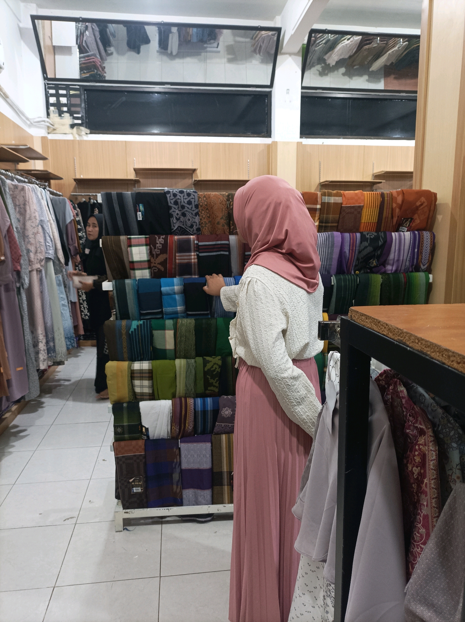 In a retail store in Blimbing, Indonesia, two women are observed. One woman, wearing a pink hijab and a textured white blouse with a pink pleated skirt, stands with her back to the camera, facing a display of folded textiles. Her right hand is extended towards the display. The second woman, dressed in a black hijab and abaya, is partially visible to the left of the frame, reaching for an item on a rack of hanging garments. The store environment features wooden shelving, tiled flooring, and displays of fabrics in various colors and patterns, including sarongs and possibly dresses. The lighting is overhead and appears to be artificial. Reflections are visible in a mirrored surface mounted high on the wall.