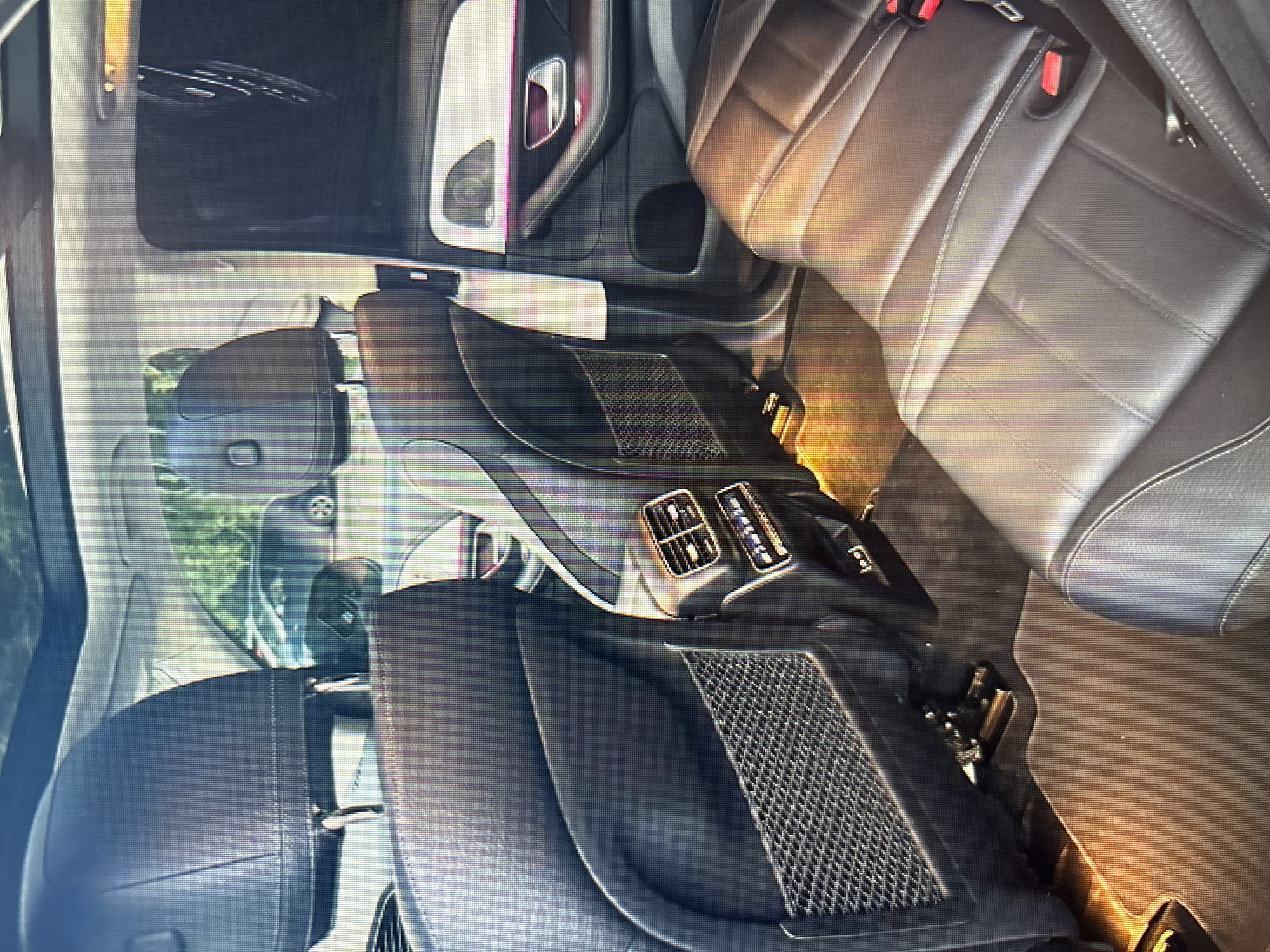 The image shows the interior of a modern car, likely a luxury SUV, with black leather seats and a panoramic sunroof. The view is from the back seat, looking forward. There are visible air vents, a door panel with ambient lighting, and seat pockets. The flooring appears to be a dark carpet. There are no people or animals visible in the image. The exterior scene visible through the windows appears to be a sunny day with trees and other vehicles in the background, suggesting an outdoor parking area. There are no specific landmarks or indicators that would pinpoint this location to Moscow, Russia.