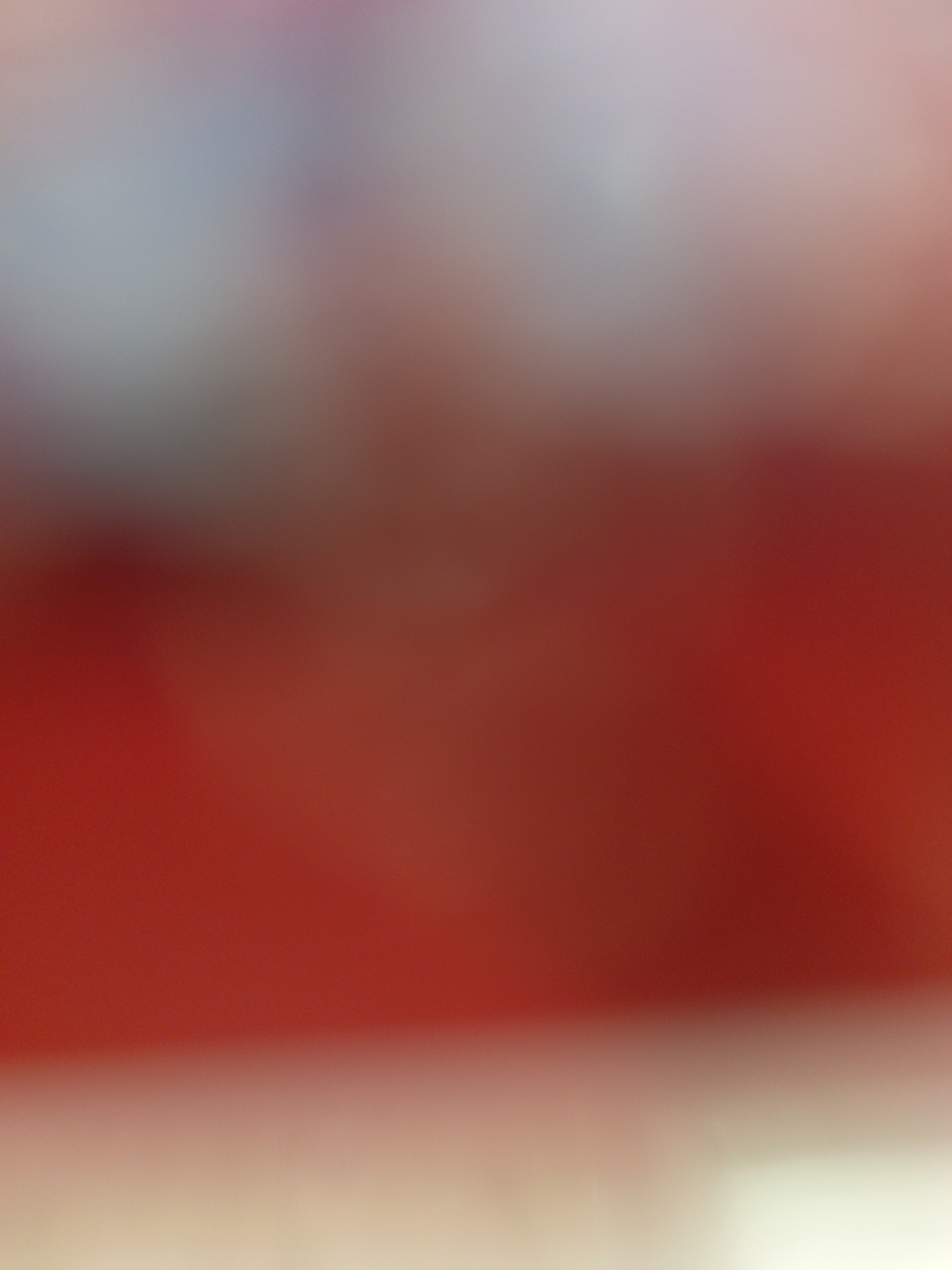 The image is a blurred, out-of-focus shot. The dominant color is red, transitioning to lighter shades of pink and white at the bottom and a hint of gray at the top. No specific objects or locations can be identified. No people, subjects, or actions are discernible.
