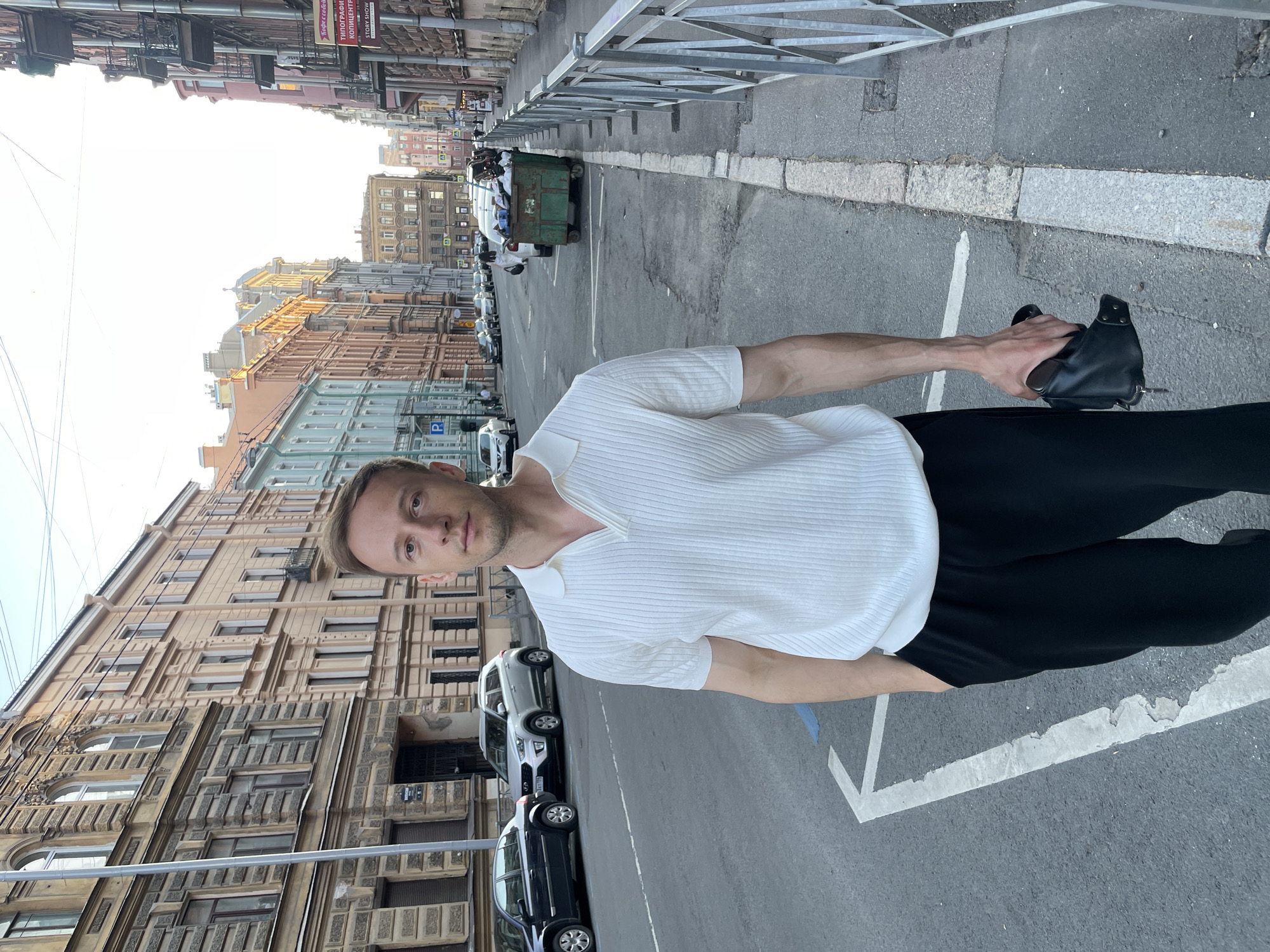 A male individual stands centered on an asphalt street in Saint Petersburg, Russia, under a bright sky, likely during late afternoon or early evening. The man, with short brown hair and light skin, wears a white ribbed short-sleeved polo shirt with an open V-neck and black loose-fitting trousers. His left hand is in his pocket, and his right hand holds a black leather pouch or wallet. He is looking directly at the viewer.
The street is flanked by multi-story, European-style buildings exhibiting ornate architectural details. On the left, buildings display varied pastel facades, including a prominent light beige building with intricate stone decorations and further structures in shades of light blue and light orange. Several cars are parked along the left curb, including a black Renault Clio and a silver Hyundai Creta. A blue "P" parking sign is visible on the sidewalk.
On the right side of the street, a metal barrier fence runs along the sidewalk, behind which another row of buildings, some with balconies, recedes into the distance. A green industrial trash dumpster, visibly overflowing with refuse, is parked beside the curb further down the street on the right. Road markings, including a prominent white arrow pointing towards the viewer, are visible on the asphalt. Cyrillic text on a commercial sign, reading "ТИПОГРАФИЯ КОПИЦЕНТР" (Printing House Copy Center) and "КОФЕ С СОБОЙ" (Coffee To Go), is visible on a building on the right side of the street, confirming the Russian location.