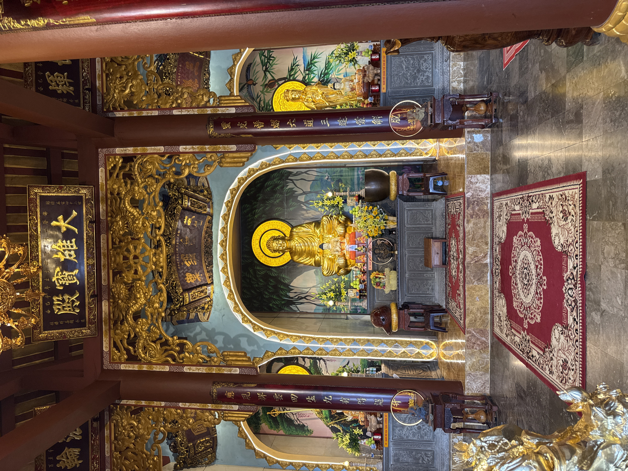 The image shows the interior of a Buddhist temple, likely in Đà Nẵng, Vietnam, given the context of the request. The central focus is a large, gilded Buddha statue seated in a meditative pose within an arched alcove. The alcove features a painted backdrop of trees and foliage. Flanking this central figure are two smaller gilded statues, one on either side, also within arched recesses.
The space is richly decorated with gold accents, intricate carvings of dragons and ornate motifs, and vibrant red carpets laid out on the marble floor. Several smaller altars and pedestals hold offerings such as flowers, fruit, and incense. Tall, dark red pillars with vertical calligraphy scripts frame the central area. The lighting is warm and appears to be a mix of natural light filtering in and artificial illumination highlighting the statues and decorative elements. The overall impression is one of opulence and reverence.