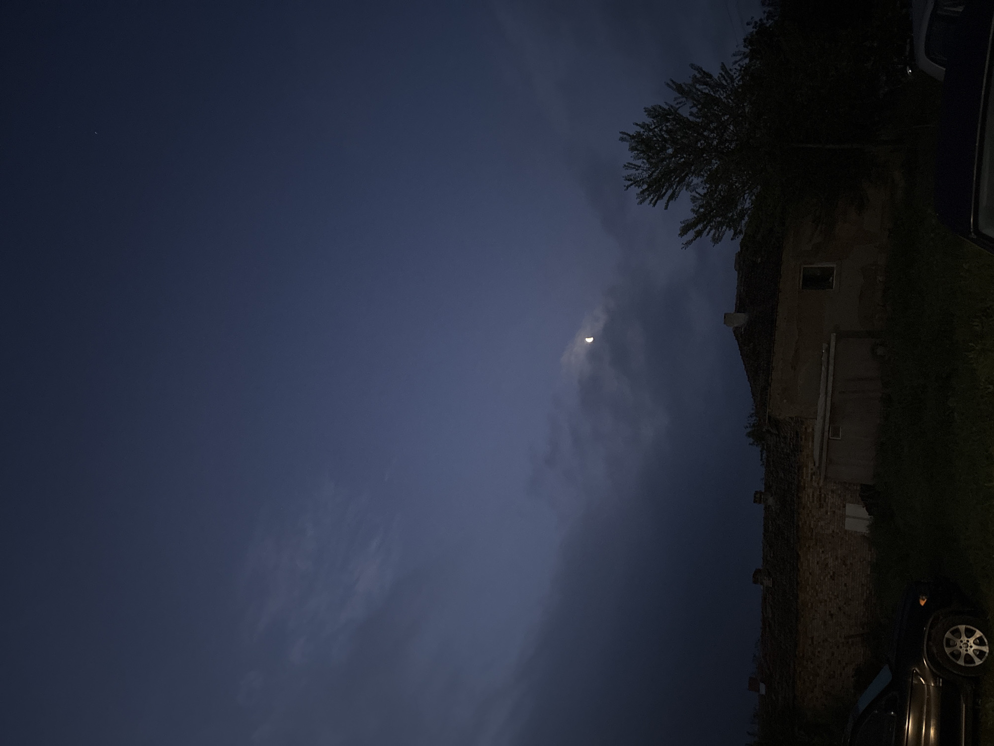 An eye-level, outdoor shot taken at night depicts a partly cloudy sky with a crescent moon, a large building with trees, and vehicles in Unknown, Unknown. The sky is dark blue with wispy clouds, and a small crescent moon is visible in the upper center. The building is to the right, it appears to be made of brick or stone. Trees and vegetation surround the building. At least one vehicle is on the right edge of the frame, with only a wheel and part of the body visible. The scene is dimly lit.