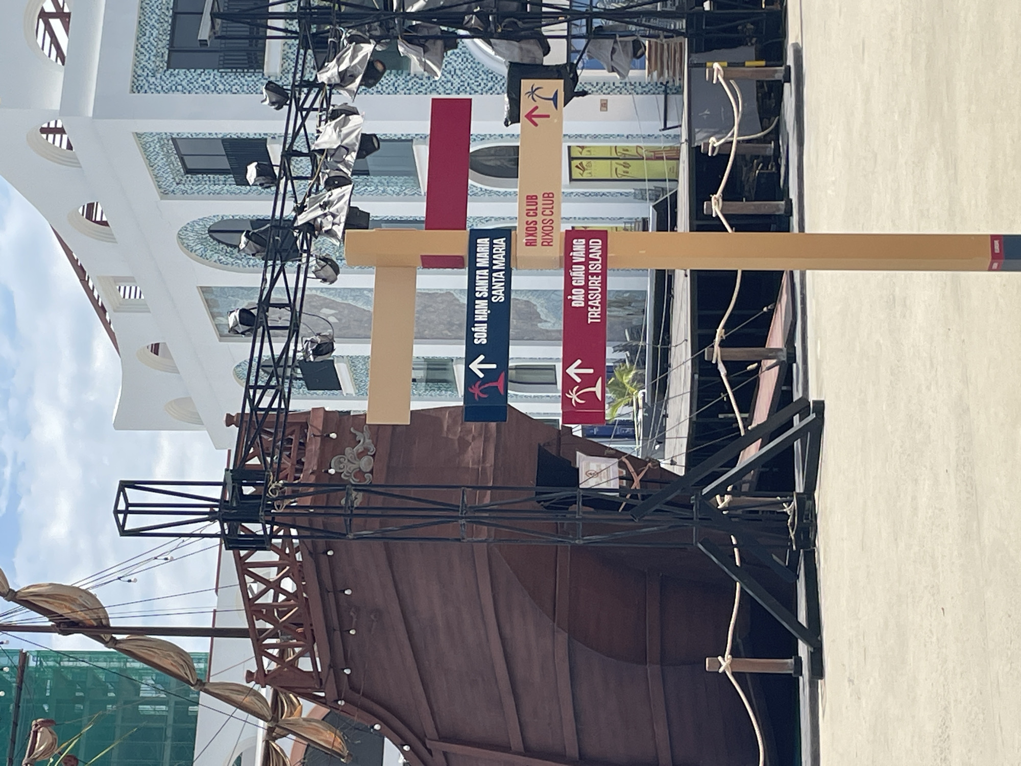 Captured in Bac Nha Trang Ward, Vietnam, the image presents a street scene featuring a complex arrangement of signage. These signs, bearing directions and names such as "SOÁI HẠM SANTA MARIA," "RIXOS CLUB," and "ĐẢO GIẤU VÀNG TREASURE ISLAND," are affixed to a framework. Overhead, a ship-themed structure or decoration is partially visible, adding a nautical theme to the urban environment. The backdrop consists of a building with arched windows, contributing to the visual complexity of the scene.