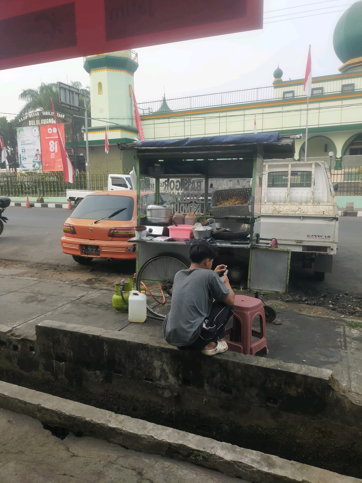 The image captures a street scene in Bululawang, Indonesia, likely near a mosque. In the foreground, a person, appearing to be male, is seated on a small red stool on a concrete ledge, looking down at a smartphone. They are wearing a gray t-shirt and dark pants with white stripes. To the left of the person, on the ledge, are two green propane tanks and a white plastic container. Behind the person is a food cart, equipped with cooking utensils and food items, attached to a white truck. A portion of an orange car is visible to the left of the food cart. The background features the distinct architecture of a mosque, including a green-domed structure and a minaret, with red and white Indonesian flags displayed. The overall environment suggests a commercial or roadside area with elements of daily life and cultural landmarks.