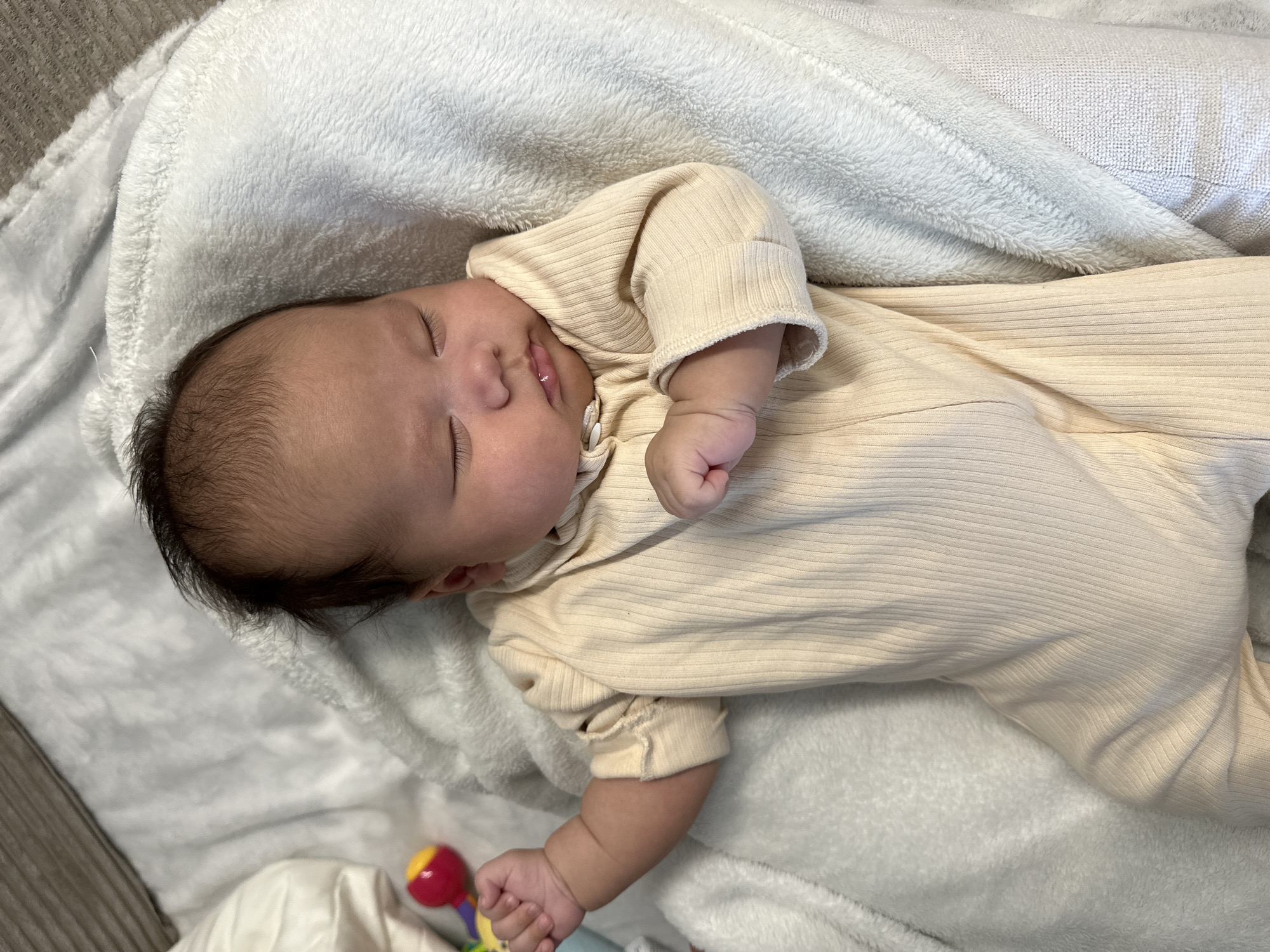 A sleeping infant lies on their back, partially turned to their right side, against a white, soft, and fluffy blanket or bedding. The baby wears a light cream or pale yellow long-sleeved, ribbed one-piece outfit. Their eyes are closed, indicating sleep, and their mouth is slightly ajar. Dark hair is visible on the baby's head. Near the baby's left hand, a small, colorful toy, possibly a rattle, with red and yellow sections, is visible. The lighting is soft and even, suggesting an indoor setting. The overall scene conveys a sense of peaceful rest. There is no visible text.