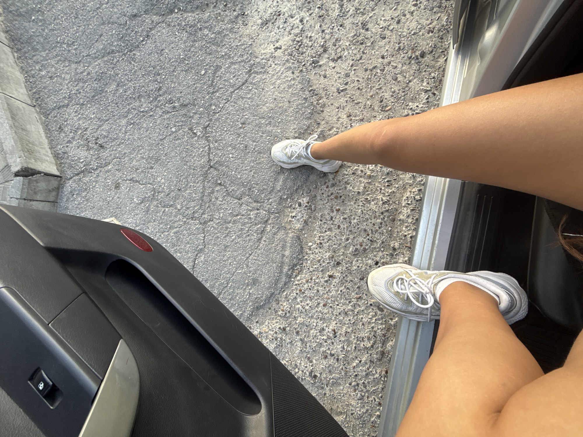The image shows a person's legs and feet, wearing white sneakers, with one leg extended out of an open car door. The car interior, with black and silver accents, is visible. The exterior ground is a rough, grey asphalt surface with visible cracks and small stones. The scene appears to be outdoors, possibly in a street or parking area. The lighting suggests daytime. The city of Cimișlia, Moldova is not visibly indicated within the image.