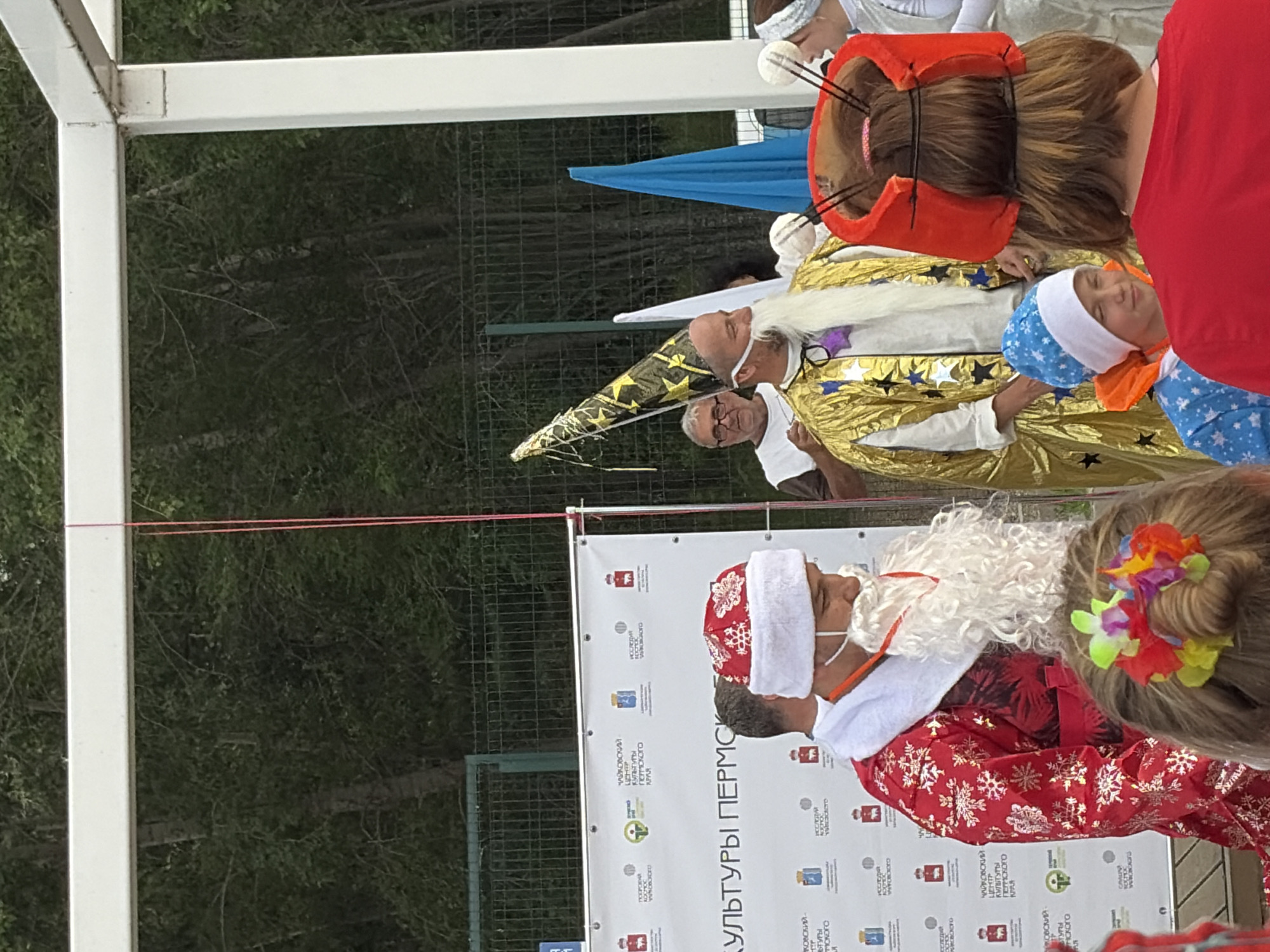 The image captures an outdoor event in Chaikovsky, Russia. In the foreground, a person dressed as Santa Claus, complete with a red suit and white beard, stands to the left. To the right of Santa, a person in a golden, star-patterned wizard's robe and a tall, conical hat with stars is visible. A banner behind them reads "КУЛЬТУРЫ ПЕРМСКОГО KPA" in large letters, indicating a cultural event. In the background, trees and a fence suggest a park or open area. Several other individuals are present, some in costume, and a blue tent is visible. The overall scene appears festive and community-oriented.