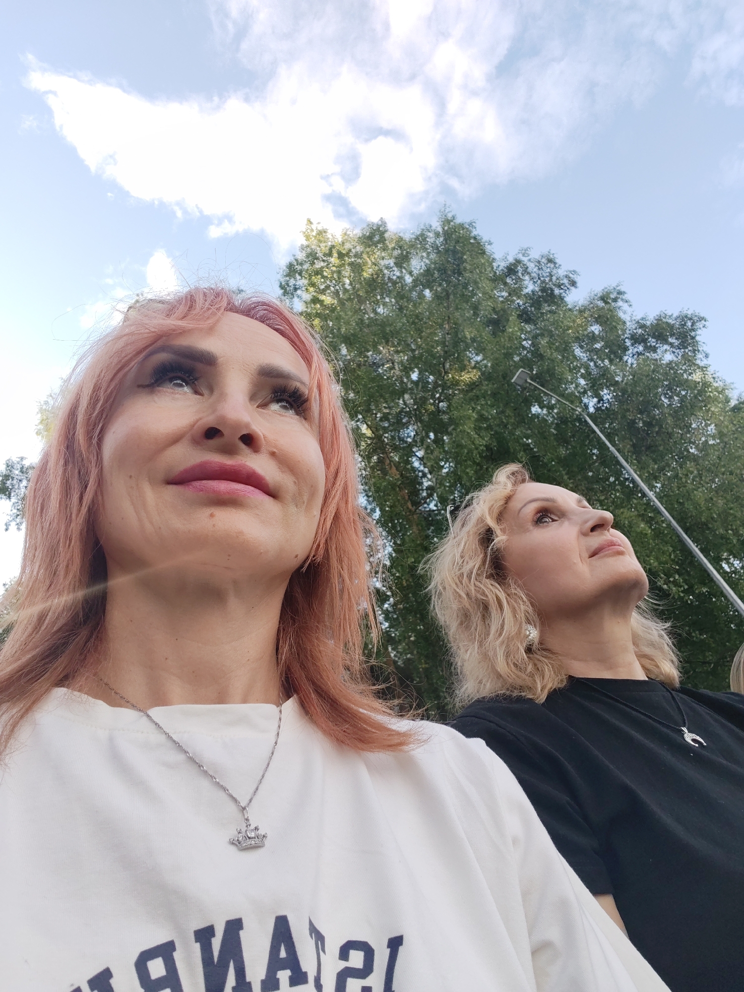 Two women are looking upwards, with trees and a blue sky with clouds in the background. The woman on the left has pink hair and is wearing a white t-shirt with a silver necklace. The woman on the right has blonde hair and is wearing a black t-shirt with a silver necklace. The scene appears to be outdoors during the daytime, under fair weather conditions. The overall emotion conveyed is one of contemplation or observation. There is some text on the white t-shirt, but it is mostly obscured.