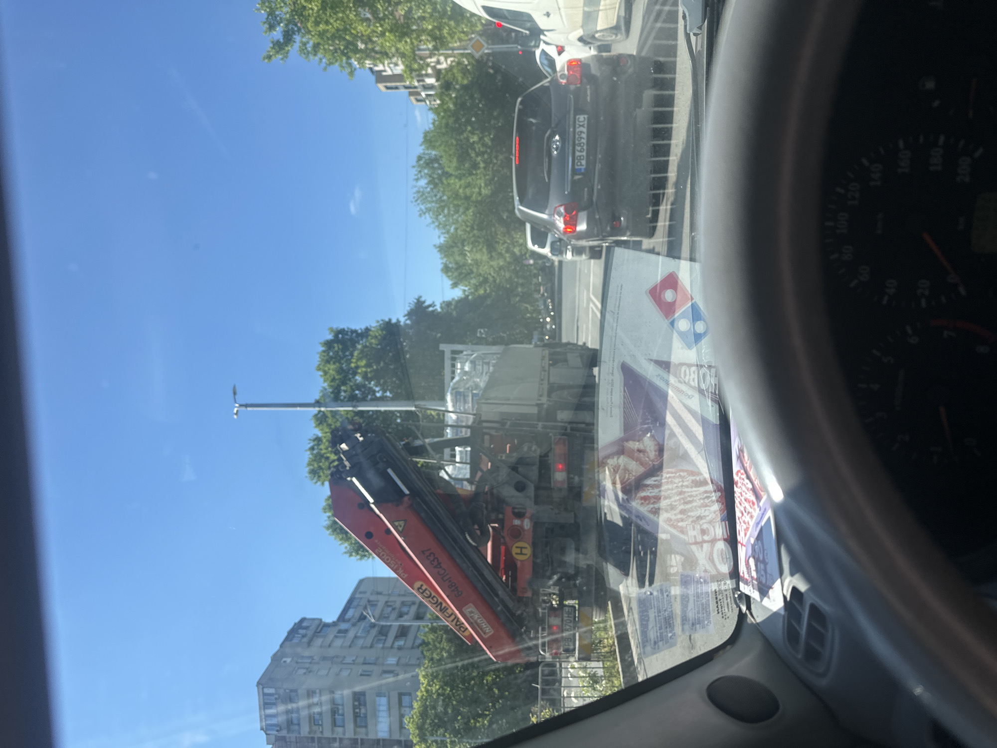 The image, taken from inside a car, shows an outdoor scene with a clear blue sky. In the immediate foreground is a section of the car's dashboard, including the speedometer, along with an advertisement for Domino's Pizza attached to the windshield.
Outside the car, various objects and vehicles are visible. There is a Palfinger crane truck with the license plate "PB 670 HE" on the right. A gray Toyota with license plate "PB 6899 XC" is also present, along with other vehicles farther in the background, including a white van.
The environment includes urban elements such as buildings and streetlights. The presence of trees and other greenery suggests an urban location, possibly in Plovdiv, Bulgaria, given the Cyrillic letters "ПС" and "PB" in the license plates of the vehicles.