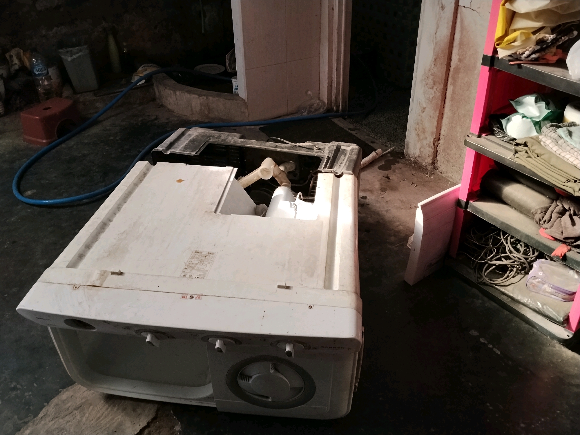In Panggungan, Indonesia, a damaged white top-loading washing machine sits on a dark, grimy concrete floor, its lid open, revealing internal components. A blue hose snakes across the floor from left to right, leading towards the machine. To the left of the washing machine, a small red stool is visible. Behind the stool, a plastic bottle of Aqua water and a white trash can are present. Further in the background, a rough-hewn concrete basin with a dark interior and a blue hose resting on its edge can be observed. A tall, off-white tiled wall with a vertical crack runs along the right side of the washing machine. To the far right, a tall pink shelving unit is partially visible, filled with various household items and coiled black cables on its shelves. The lighting is dim and uneven, casting strong shadows.