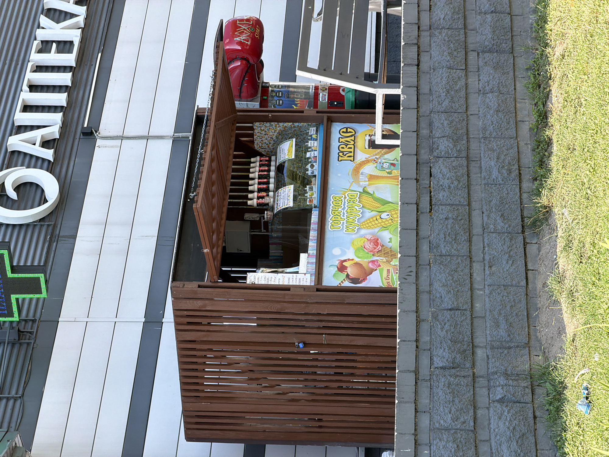 The image captures an outdoor kiosk selling ice cream and kvass, a traditional Slavic fermented beverage. The kiosk is situated in front of a building with a corrugated metal facade, identifiable by the large white text on the building that reads "АПТЕКА" (Apteka), meaning "pharmacy" in Russian. Above the kiosk, a green pharmacy cross symbol is visible. The kiosk itself is constructed of wood and features a glass display case showcasing various ice cream flavors and beverages. A colorful mural on the front of the kiosk depicts corn, ice cream cones, and glasses of kvass, with Russian text that translates to "Hot Corn" and "Kvass". A pair of red boxing gloves, emblazoned with the word "BOXE", hang above the display, suggesting a possible novelty attraction or game associated with the establishment. The kiosk is protected by a wooden slatted gate, which is secured with a blue padlock. The scene is set against a backdrop of a gray brick wall and a grassy area in the foreground. There are no people visible in the image. This type of outdoor kiosk is a common sight in urban areas, particularly in Eastern European cities like Minsk, Belarus, where they serve as convenient points for purchasing refreshments and snacks.