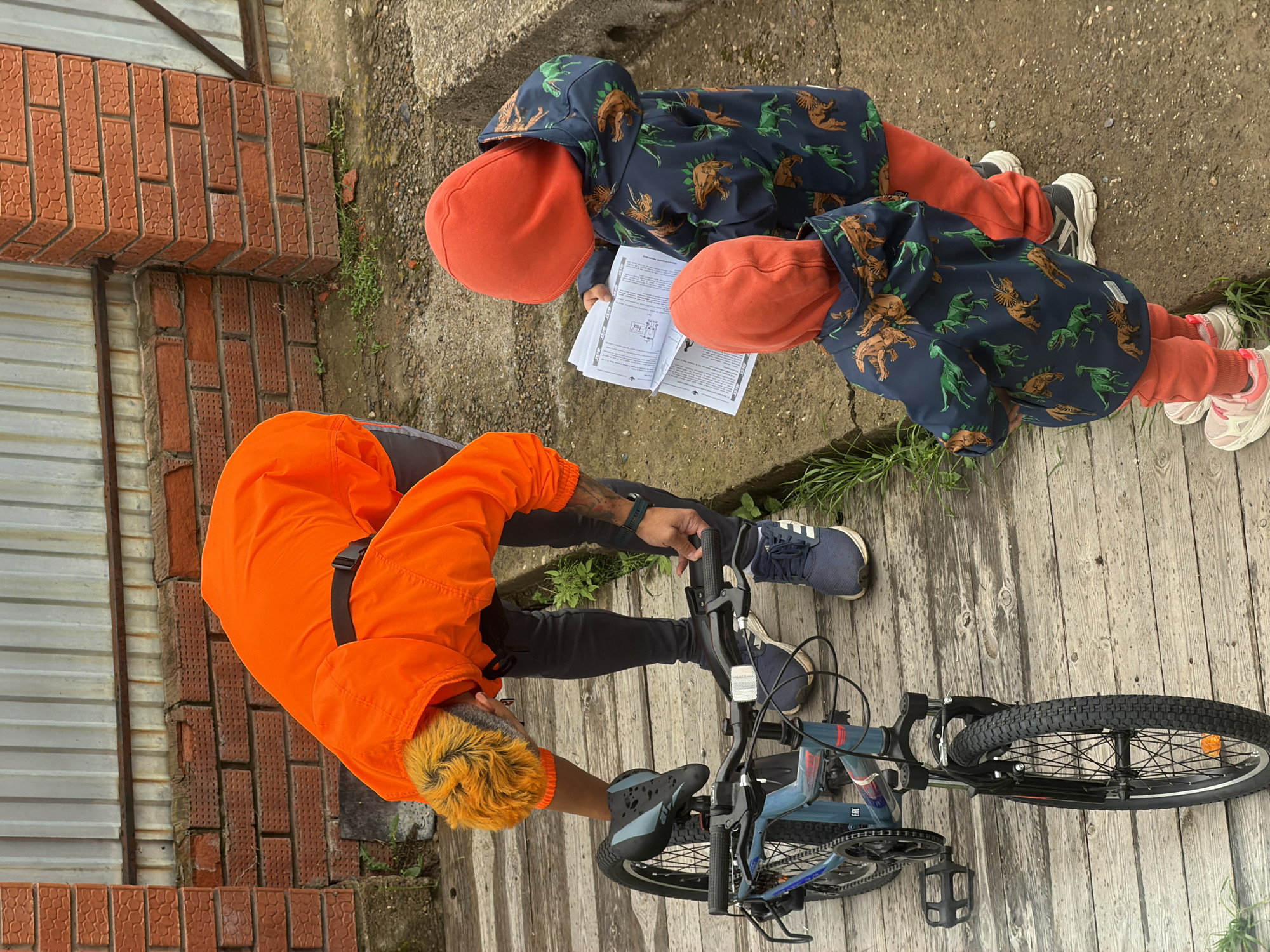 The media file shows an outdoor scene with three people and a bicycle. Two children, wearing hooded jackets with a dinosaur print and orange hats, are looking at a booklet. One child is sitting, and the other is partially visible behind them. A person with vibrant orange hair, wearing an orange jacket, is kneeling and appears to be adjusting the bicycle. The bicycle is blue and black.
The setting appears to be a weathered wooden boardwalk next to a brick wall and some greenery. The ground is made of wooden planks, and there is a concrete area to the right. The lighting suggests it is daytime, possibly overcast.
There is no visible text in the media file. The overall mood seems casual and perhaps educational, as the children are engaged with the booklet. The location cues suggest an outdoor area, possibly a backyard or a public walkway.