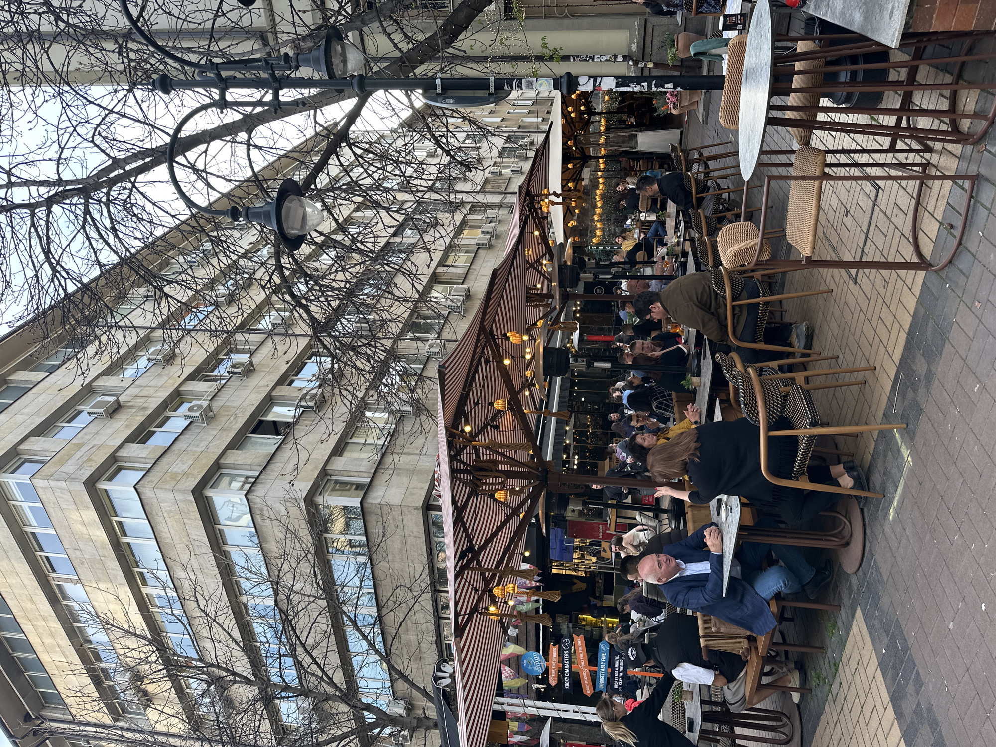 The image shows an outdoor cafe scene in Sofia, Bulgaria. The cafe is situated on a sidewalk with tables and chairs occupied by patrons. A large building looms in the background, its facade marked by numerous windows and air conditioning units. A bare tree with many branches is visible on the left side of the frame. A streetlamp is positioned near the tree. The cafe's awning provides shade and decorative lighting. Advertisements are affixed near the entrance of the cafe. The sidewalk is paved with bricks.