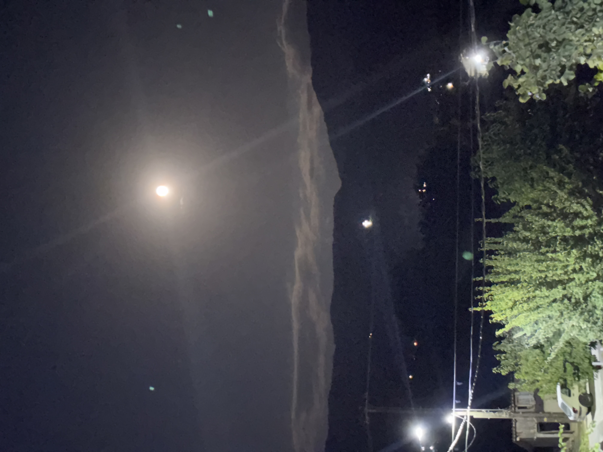 A night-time photograph captured from an elevated position overlooks the city of Leskoec, North Macedonia. The sky is dark with a bright, hazy moon positioned in the upper center. Streaks of light, likely from lens flare, emanate from the moon and other light sources. Below the moon, horizontal bands of clouds are visible against a dark mountainous landscape. The foreground features power lines stretching across the frame, supported by utility poles. Some distant lights, possibly from buildings, are scattered across the hillsides. In the lower portion of the image, the branches of trees and shrubbery are illuminated by artificial light. A small white car is parked on the left side of the frame.