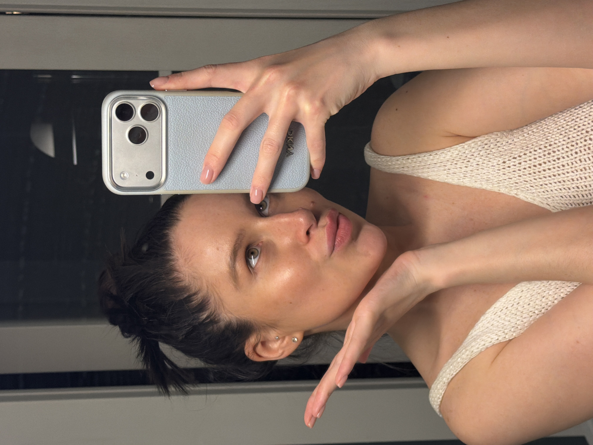 The image shows a mirror selfie taken by a young woman in what appears to be an indoor setting in Khimki, Russia. The image is rotated 90 degrees counter-clockwise.
The woman is the central subject, holding a light blue or grey textured smartphone case with her left hand to take the photo. Her right hand is raised gracefully towards her face, with fingers slightly fanned. She has dark hair, styled perhaps in a ponytail or bun, and her light brown or hazel eyes are directed towards the phone's camera. Her expression is a subtle pout, lending a slightly playful or contemplative mood. She wears minimal makeup, highlighting natural features, including visible freckles. Two small stud earrings are visible on her left earlobe.
She is dressed in a light-colored, possibly cream or off-white, knitted or crocheted top with a noticeable open weave texture, worn off one shoulder. The background is a dark, reflective surface, indicating a mirror, with faint horizontal lines suggesting an interior space like a bathroom or a hallway.
Notable details include the smartphone, which features a prominent silver-toned camera module housing three lenses, a flash, and another sensor. The text "DKNY" is visible vertically aligned on the light blue phone case. The lighting appears artificial and even, typical of an indoor setting, without strong external light cues to suggest the time of day beyond being indoors.