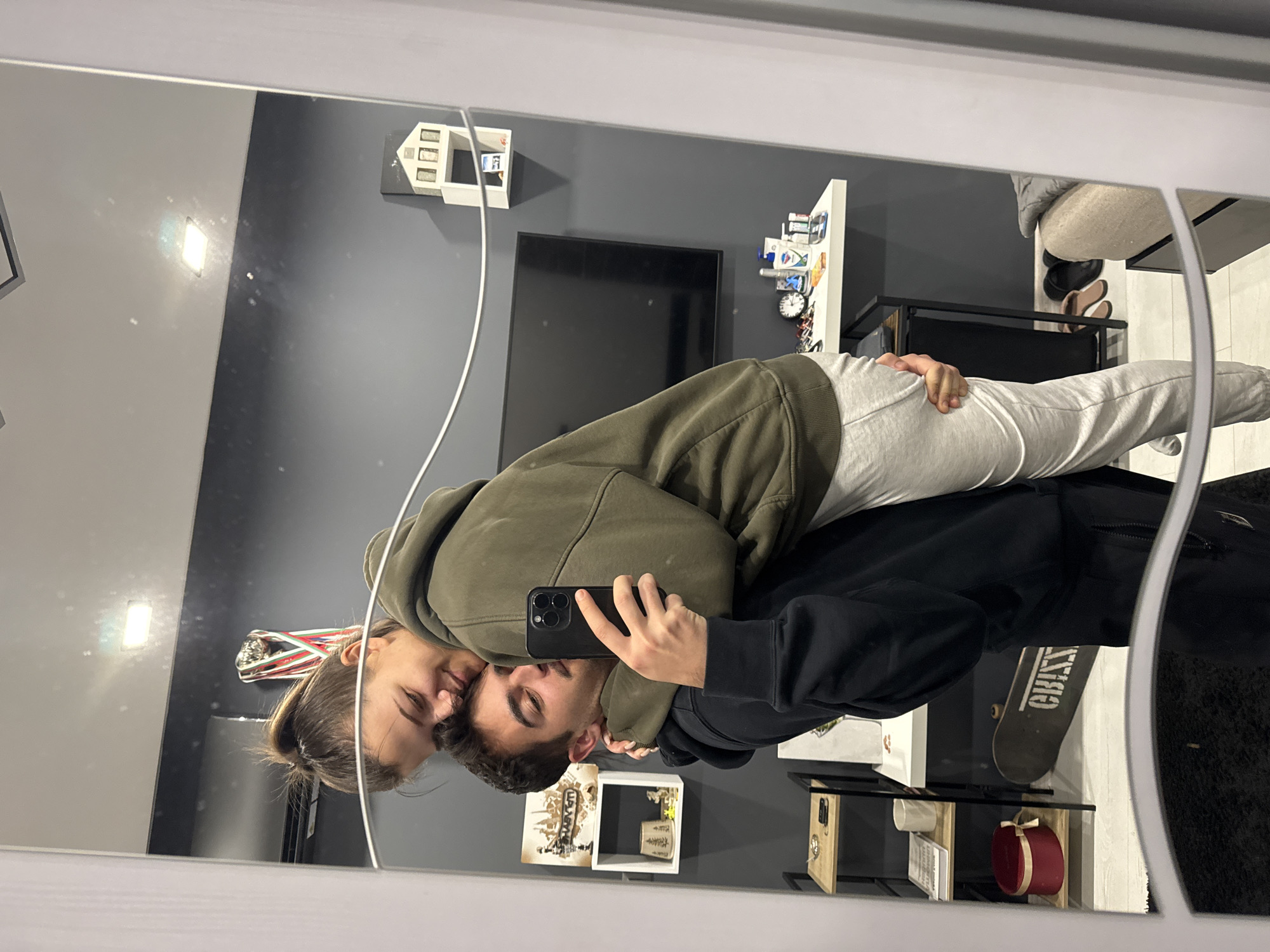 A mirrored reflection shows a young couple taking a selfie in what appears to be an indoor setting. A young woman with her hair in a bun wears a green hoodie and embraces a young man who holds a smartphone. The man wears a black hoodie. Behind them is a wall painted a dark grey color. A flat-screen television is mounted on the wall, and a decorative white house-shaped shelf is above it. To the right of the television is a white shelf unit with various items on display. A black metal shelf unit with a glass top, also containing miscellaneous items, is beneath the couple. A skateboard is leaning against the side of this shelf. The room has a casual and lived-in feel. It is not possible to determine if this image was taken in Sofia, Bulgaria.