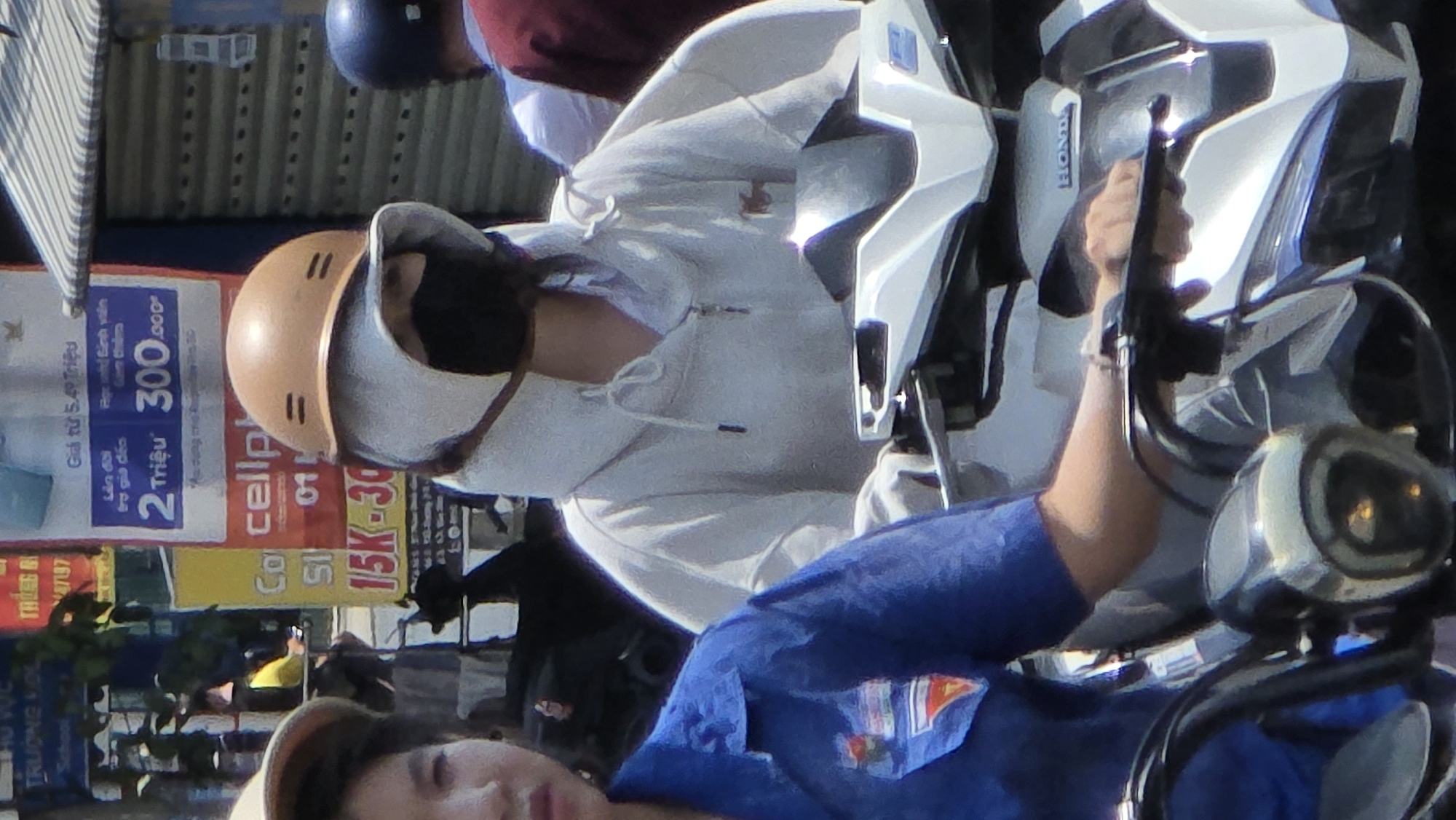 Captured in Bắc Nha Trang, Vietnam, the image presents a street scene dominated by two individuals on a motorbike. One, wearing a tan helmet and a light-colored hooded sweatshirt paired with a face mask, sits on a white Honda scooter. A second person, wearing a blue collared shirt, faces the camera in the foreground. The motorbike is prominent, its controls and mirrors in clear view. Backdrops feature street signage including cell phone advertisements.