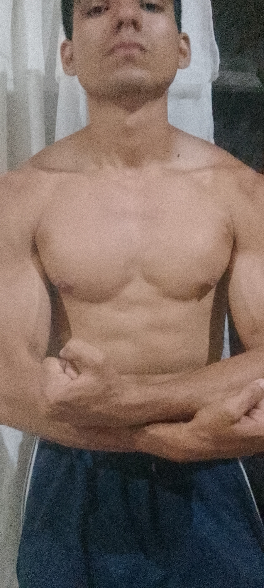 A torso of a man is seen in the foreground. He has a light complexion, dark hair, and is shirtless. His arms are folded across his abdomen. He is wearing dark blue shorts with a white stripe on the side. In the background, there is a white curtain, and further back, a dark, unidentifiable space. The location cannot be identified from the information in the image.