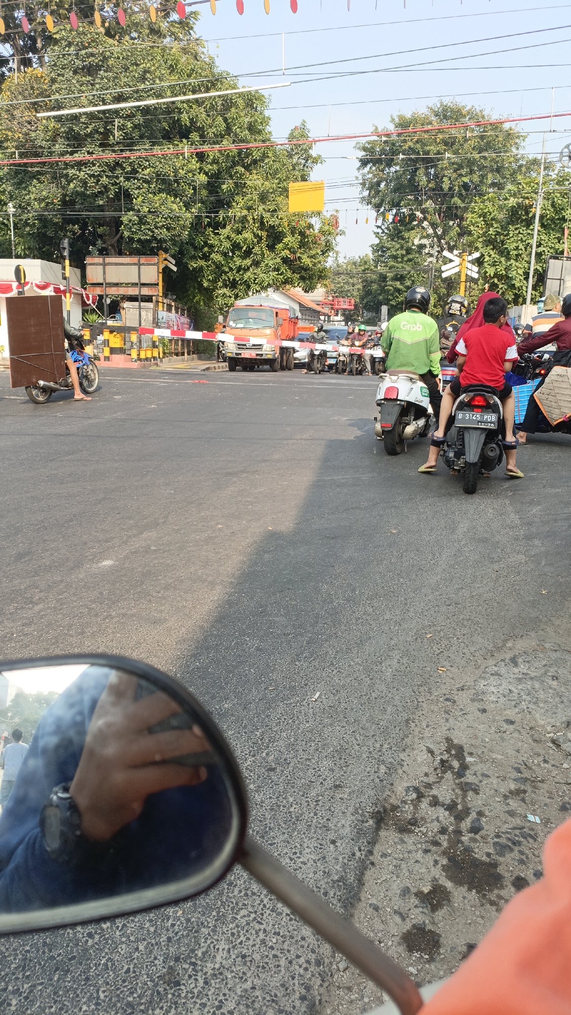 The image depicts a traffic scene in Central Jakarta, Indonesia. In the foreground, a motorcycle's mirror reflects a hand wearing a watch and a blue long-sleeved shirt. The road surface is asphalt. Beyond the mirror, a group of motorcycles are stopped at what appears to be a barrier, possibly a toll gate or checkpoint.
Several individuals are visible on motorcycles. One person in a green jacket with "Grab" branding is on a white scooter. Another motorcycle has two people on it, one wearing a red shirt and the other a red shirt with white trim. There are other motorcycles and people further back in the queue.
A red truck is positioned in the middle of the road, with its hazard lights on. Barriers, some with red and white stripes, are set up across the road. In the background, there are lush green trees and overhead wires. The sky is a clear, light blue. Decorative colorful flags are strung across the top of the image. The overall impression is of a busy urban intersection with traffic congestion.
