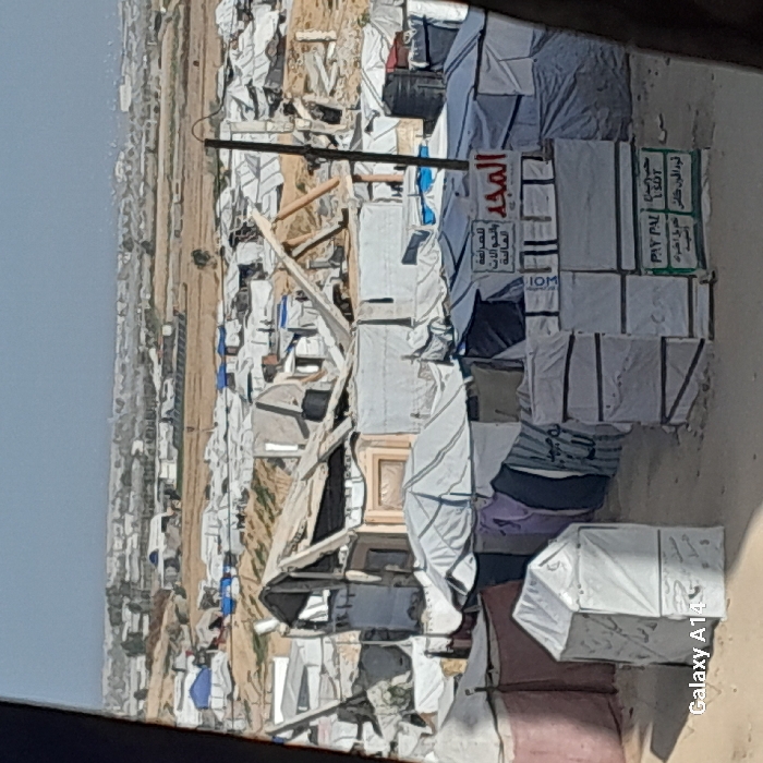 A camp of tightly packed tents dominates the frame, with the tents' white fabric contrasting against the surrounding sandy terrain. A sign with Arabic script is prominently displayed amidst the tents. The overall scene depicts a dense and likely temporary settlement, possibly a refugee camp or a similar emergency housing situation. The presence of the sign and organized structures suggests a degree of planning within the camp.