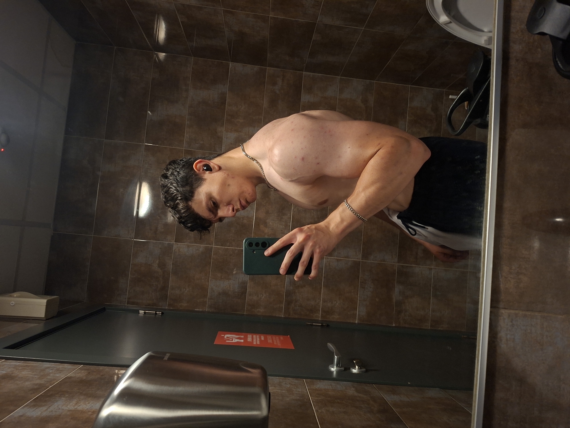 This media file shows a young man taking a mirror selfie in a public restroom, likely in Plovdiv, Bulgaria. He is shirtless, wearing dark shorts and a silver chain necklace, with black earbuds in his ears. His posture suggests he is flexing or posing, showcasing his physique. He is holding a dark green smartphone, which he is using to capture the image.
The restroom appears to be tiled with brown ceramic tiles on the walls and floor. Visible in the reflection are a toilet tank, a sink, and a door with a red sign that has some text in an unknown language. There is also a metallic hand dryer visible in the foreground, partially obscuring the lower part of the frame. The lighting is artificial, typical of an indoor setting. The overall mood is casual, as the man is documenting himself in a commonplace location. There are no clear indicators of the time of day or weather conditions outside.