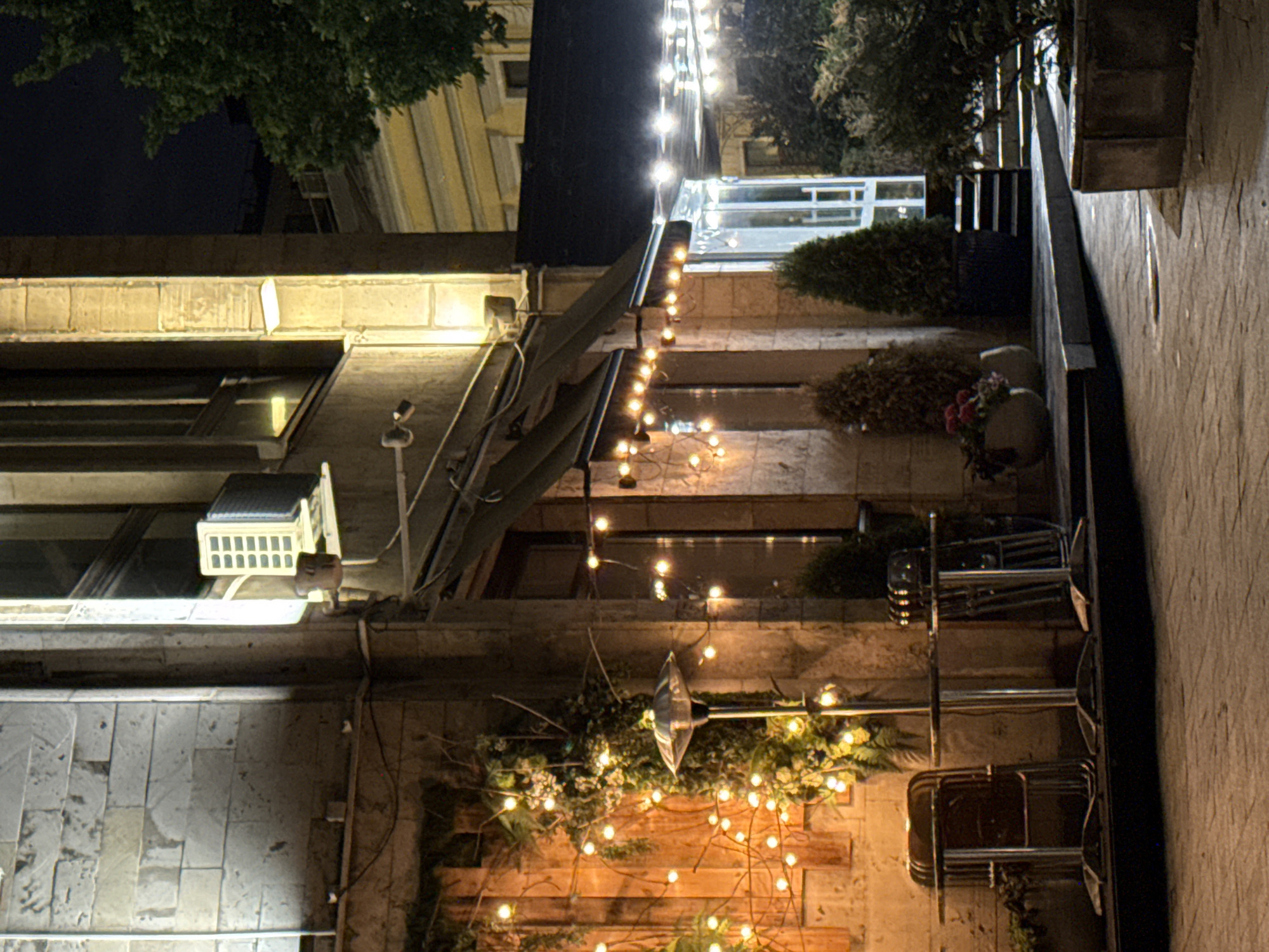 The image captures an exterior nighttime view in Moscow, Russia, featuring building facades and outdoor lighting. A row of strung fairy lights stretches across the scene, providing illumination and adding a festive ambiance. The building exteriors exhibit varying architectural styles and materials, including brick and stone. Foliage and decorative plants are present, enhancing the aesthetic appeal. Several stacked chairs are visible, hinting at an outdoor seating area. Other distinct features include window awnings, lighting fixtures, and an air conditioning unit mounted on one of the buildings.