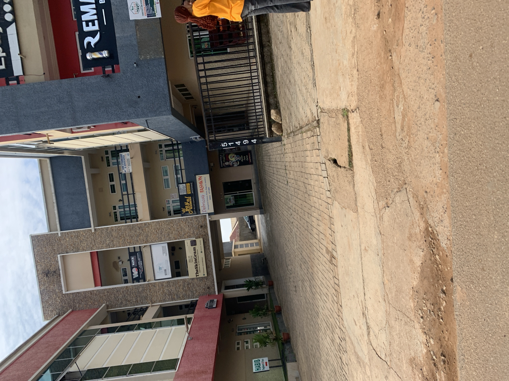 The image shows a street scene in Gura Top, Nigeria. A multi-story building with a red and cream facade is visible on the left, while a grey and blue building is on the right. Several signs are attached to the buildings, including "TheSkinGram," "Debricas FASHION," and a sign with the number "51494." A person in an orange shirt and grey pants is standing near the right side of the frame. The ground is paved with interlocking bricks and concrete, with some areas appearing worn and damaged. Potted plants are placed near the building entrances. The sky is overcast.