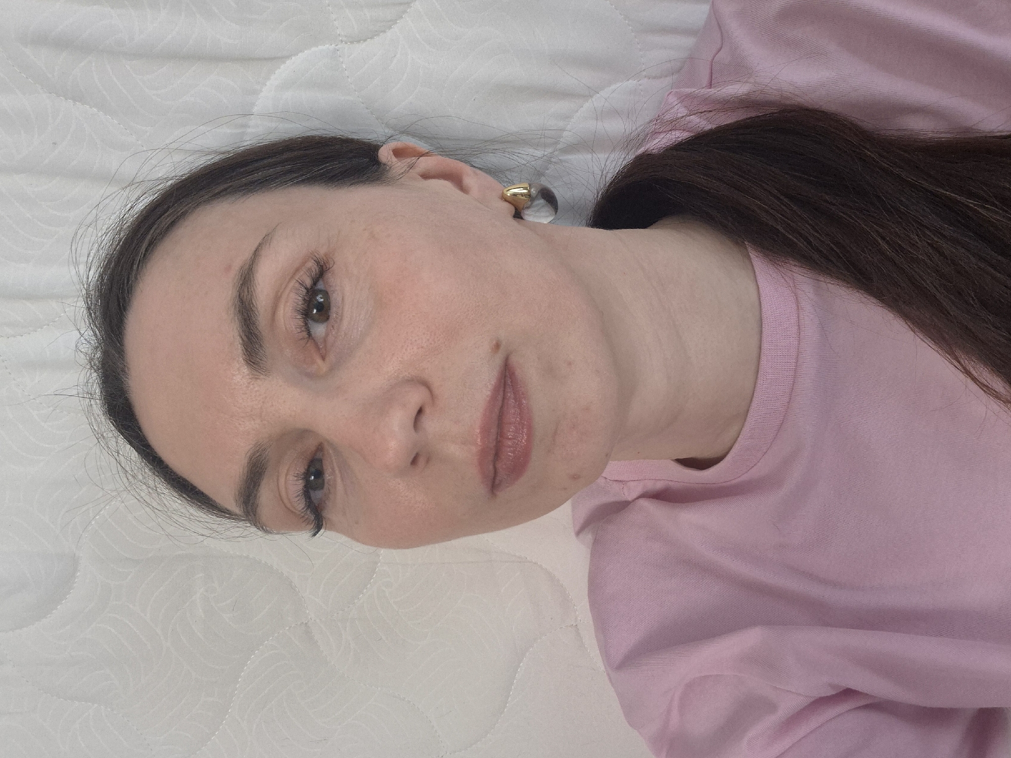 A woman lies on a white, quilted surface, possibly a bed, wearing a pink t-shirt. She has brown hair, and a clear and gold earring in her left ear. Location: Unknown, Unknown.