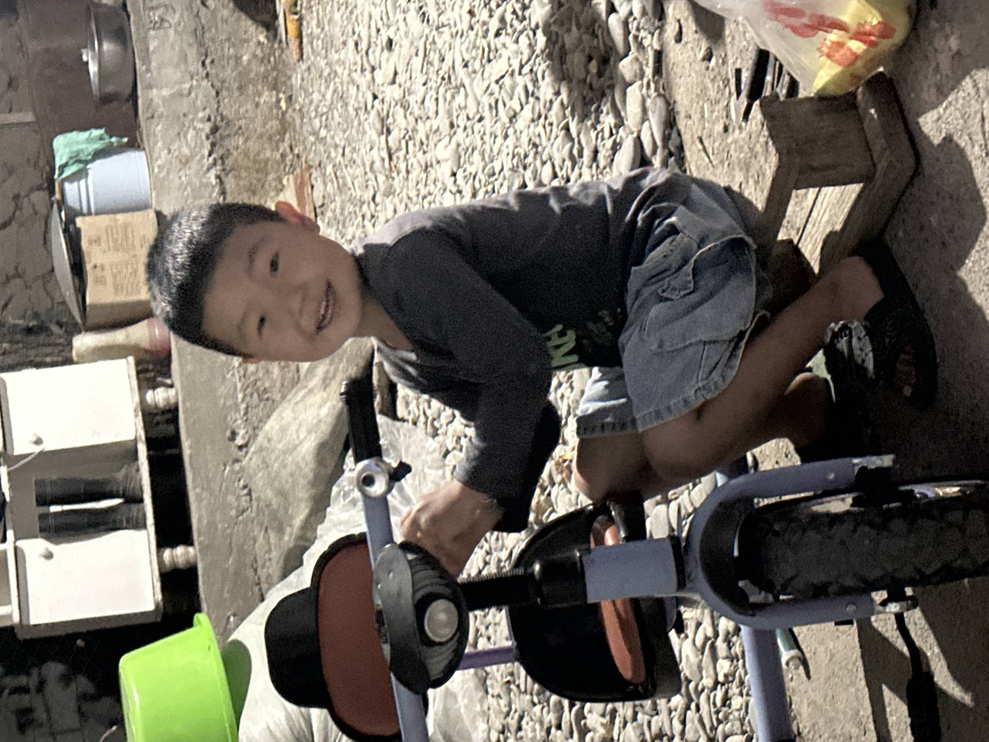 A young boy with black hair and a wide smile is sitting on a wooden stool next to a blue and purple tricycle. He is wearing a dark gray long-sleeved shirt and light blue denim shorts. The tricycle has a red seat and a black handlebar with a bell. The boy's right hand is on the handlebar. The ground is covered with small gray and beige stones. In the background, there is a white cabinet with two black boots inside. To the left of the cabinet, a bright green plastic basin is visible. Further in the background, there are buckets, boxes, and a metal bowl. The scene appears to be outdoors, possibly in a yard or alleyway. The lighting suggests it is daytime with sunlight casting shadows. The overall environment seems rustic and utilitarian. The image was taken in the city of Taygarayev, Kyrgyzstan.