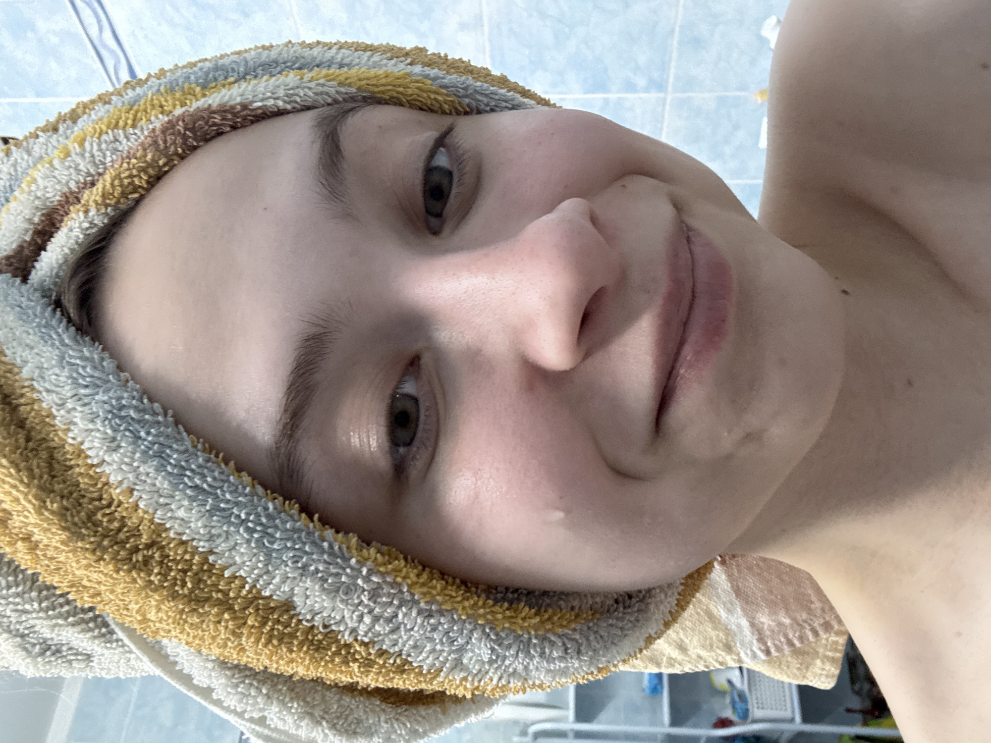 A close-up, selfie-style photograph depicts a young woman with a towel wrapped around her head, likely after a shower. Her skin appears moist, with a visible water droplet on her cheek. The background shows a bathroom setting with blue tiles. The photograph gives no indication of being taken in Прикамский, Russia.