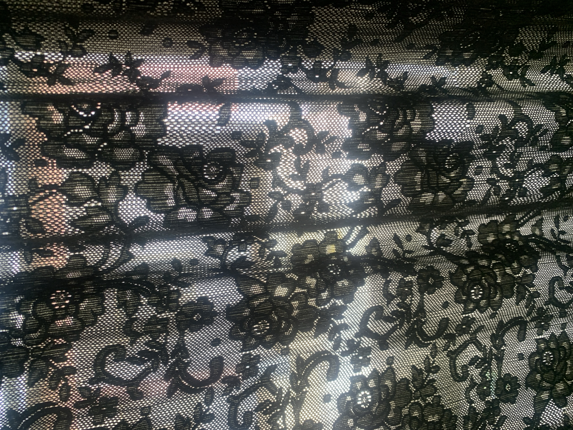 The image is a close-up, full-frame shot of a black lace curtain. The curtain has a floral pattern with intricate details. The lace is sheer, allowing light to pass through, creating a mottled effect with bright areas and darker shadows. The background visible through the curtain appears to be a wall with a textured surface and possibly some warm-toned colors, suggesting it might be illuminated by natural light from a window. The folds and draping of the fabric create a sense of depth and texture. No people, specific objects, or actions are discernible, as the focus is entirely on the textile. The scene is indoors, with the light source external to the frame. The image lacks any context of a specific location within Jauro Yinu, Nigeria, as it only depicts a curtain.