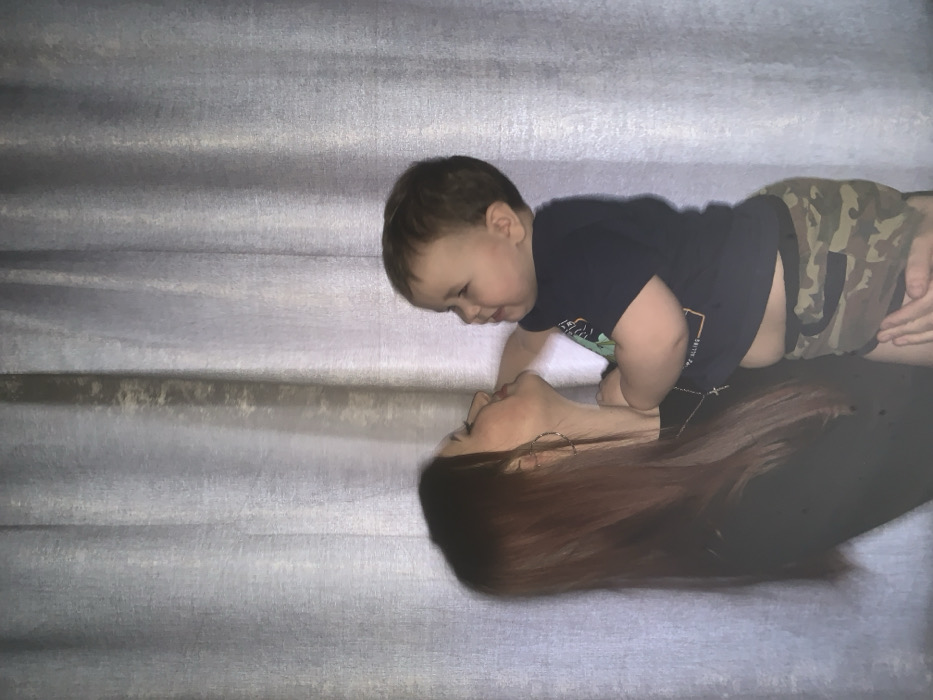 An indoor scene depicts a woman and a young child in the city of Фоки, Russia, against a backdrop of light-colored, vertically folded curtains. The woman, positioned on the left, has long, reddish-brown hair, wears a dark top, a silver-colored cross pendant, and a hoop earring on her visible left ear. She faces right in profile, with a slight smile, looking at the child. She supports the child with her visible left hand. The child, a toddler with short, dark brown hair, is held by the woman and positioned on the right, facing left towards her. The child wears a dark blue t-shirt with a partially visible light graphic and camouflage-patterned shorts. The child's left hand is raised towards the woman's face, near her cheek or chin. Both individuals are engaged in eye contact.