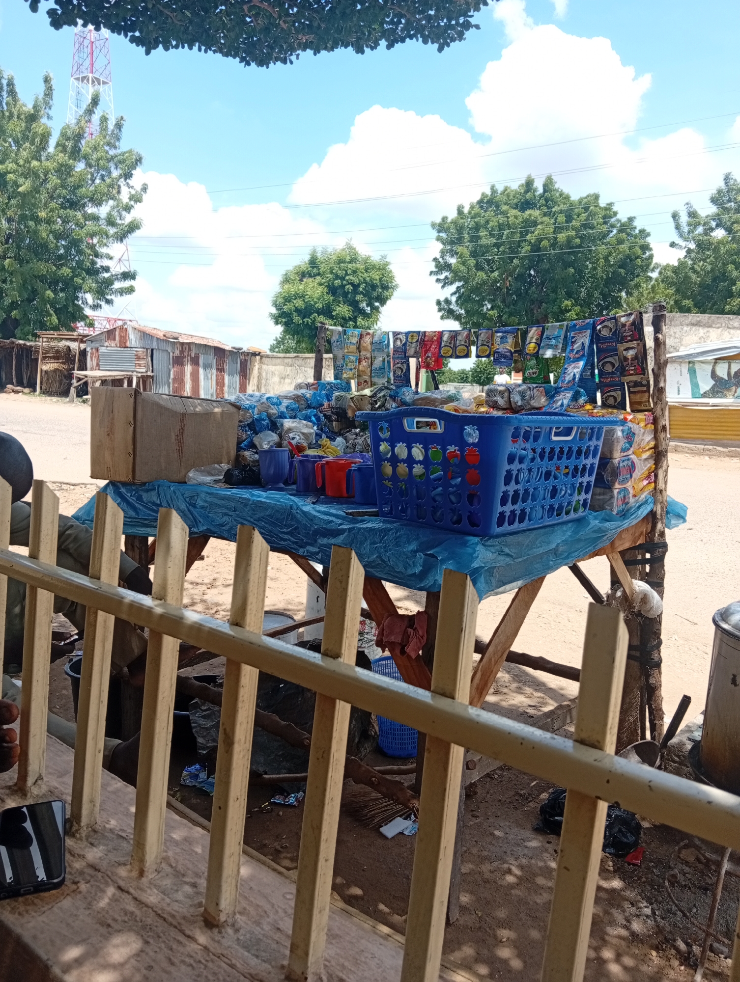 The image depicts an outdoor scene at a market stall in Zongoma Kyari, Nigeria. A wooden table, draped with a blue plastic sheet, displays various items for sale. Prominently featured are several blue plastic cups and two red plastic cups, a large blue plastic basket filled with what appears to be packaged goods, and a cardboard box. Behind the table, a rack holds numerous small, colorful packets of merchandise, possibly snacks or food items. In the background, there are corrugated metal structures, trees, and a distant telecommunications tower. The ground is sandy, and the sky is blue with scattered clouds. A person's arm is partially visible on the left side of the image, suggesting their presence at or near the stall. A mobile phone lies on a ledge in the foreground, and a metal railing partially obstructs the view.