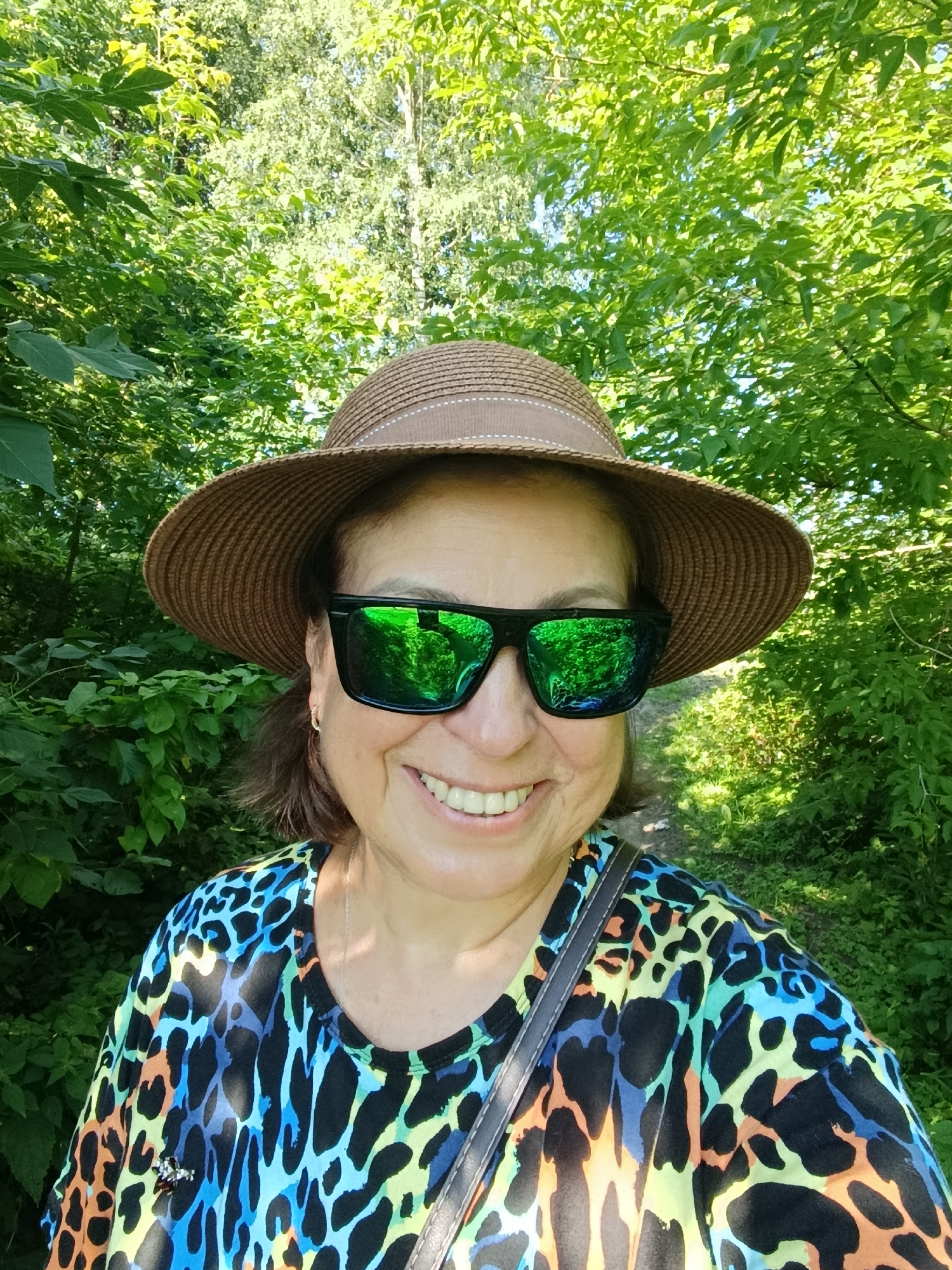 A woman with brown hair, wearing a straw hat, bright patterned t-shirt, and green mirrored sunglasses, smiles at the camera. The background consists of lush green foliage, suggesting an outdoor, possibly park-like setting. The image does not contain enough information to determine the specific location within the city of Vereya, Russia.