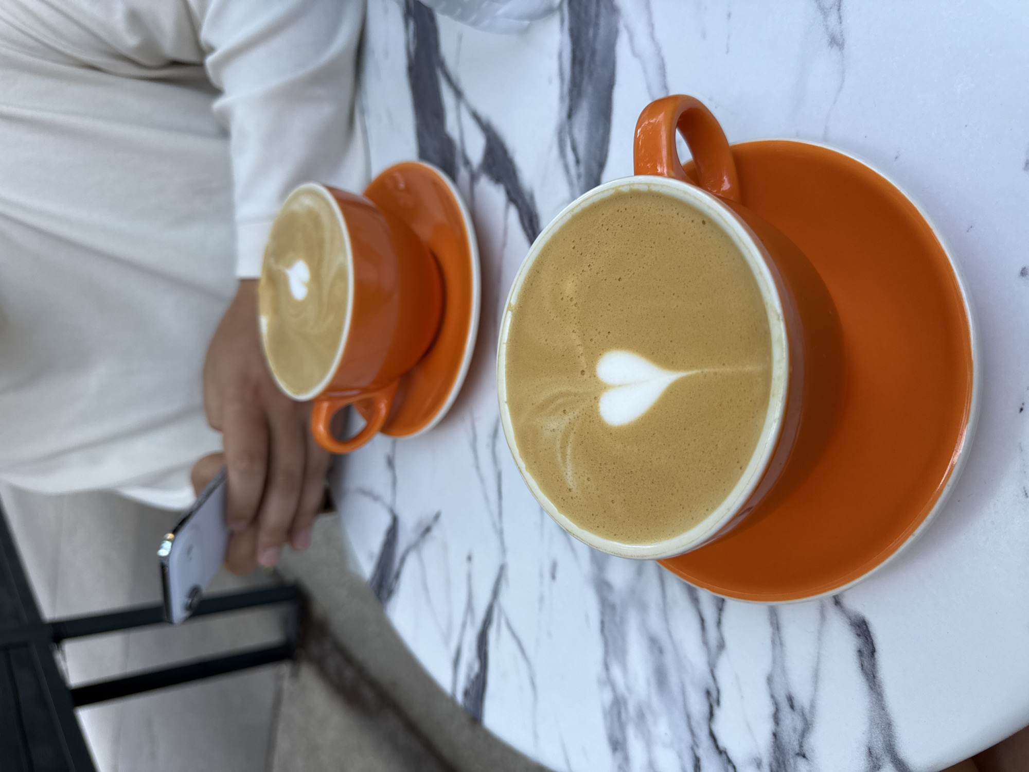 This media file captures a close-up, overhead view of two orange ceramic cups of coffee with latte art, placed on a marble surface. The coffee in the foreground cup displays a heart-shaped latte art design. The cups and their saucers are vibrant orange, contrasting with the white and gray veining of the marble tabletop. A person's hands are visible in the frame, one holding a silver smartphone. The person is wearing a white garment, possibly a long-sleeved shirt or robe. The scene appears to be set in a cafe or a similar relaxed setting, possibly outdoors given the hint of a dark railing in the lower left corner. The lighting suggests daytime. The overall mood is calm and leisurely, suggesting a moment of enjoying coffee. No visible text is present in the image.