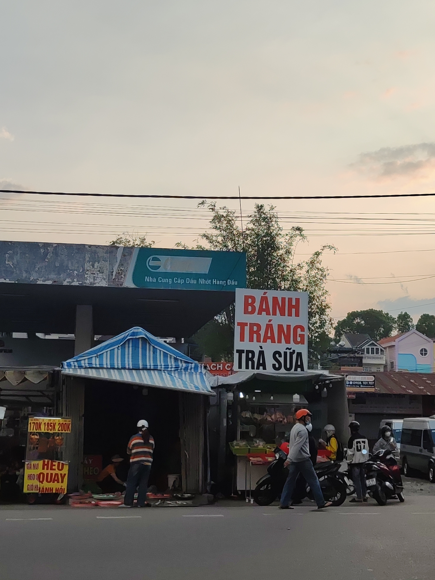 The image shows a street scene in Da Lat, Vietnam, featuring small roadside businesses. A gas station is visible in the background, with a sign that reads "Nhà Cung Cấp Dầu Nhớt Hàng Đầu." To the left, a small food stall with a blue and white striped awning sells roast pork ("Heo Quay"), the menu written on a sign in Vietnamese. A person wearing a white helmet and striped shirt stands near the stall. To the right of the food stall is a "Banh Trang Tra Sua" (rice paper and milk tea) vendor, as indicated by a large sign. Several people are standing near parked motorbikes, some wearing helmets and face masks. Overhead, power lines stretch across the sky. The sky is a pale orange due to either sunrise or sunset.