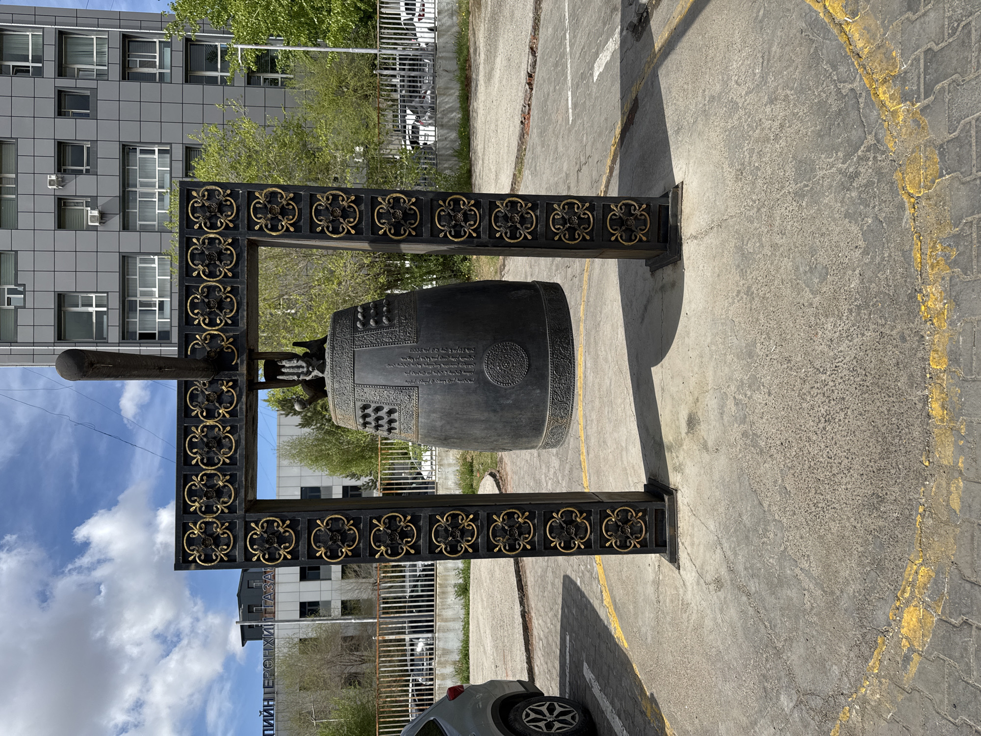 The image shows a large, ornate metal bell mounted on a black, decorated frame. The frame is positioned on a paved surface with painted yellow lines. Behind the bell, a modern, multi-story building with a gray tiled exterior is visible. A tree with green leaves is partially obscured by the building. In the foreground, a portion of a gray car is visible. The scene appears to be in an urban setting, specifically Ulaanbaatar, Mongolia.